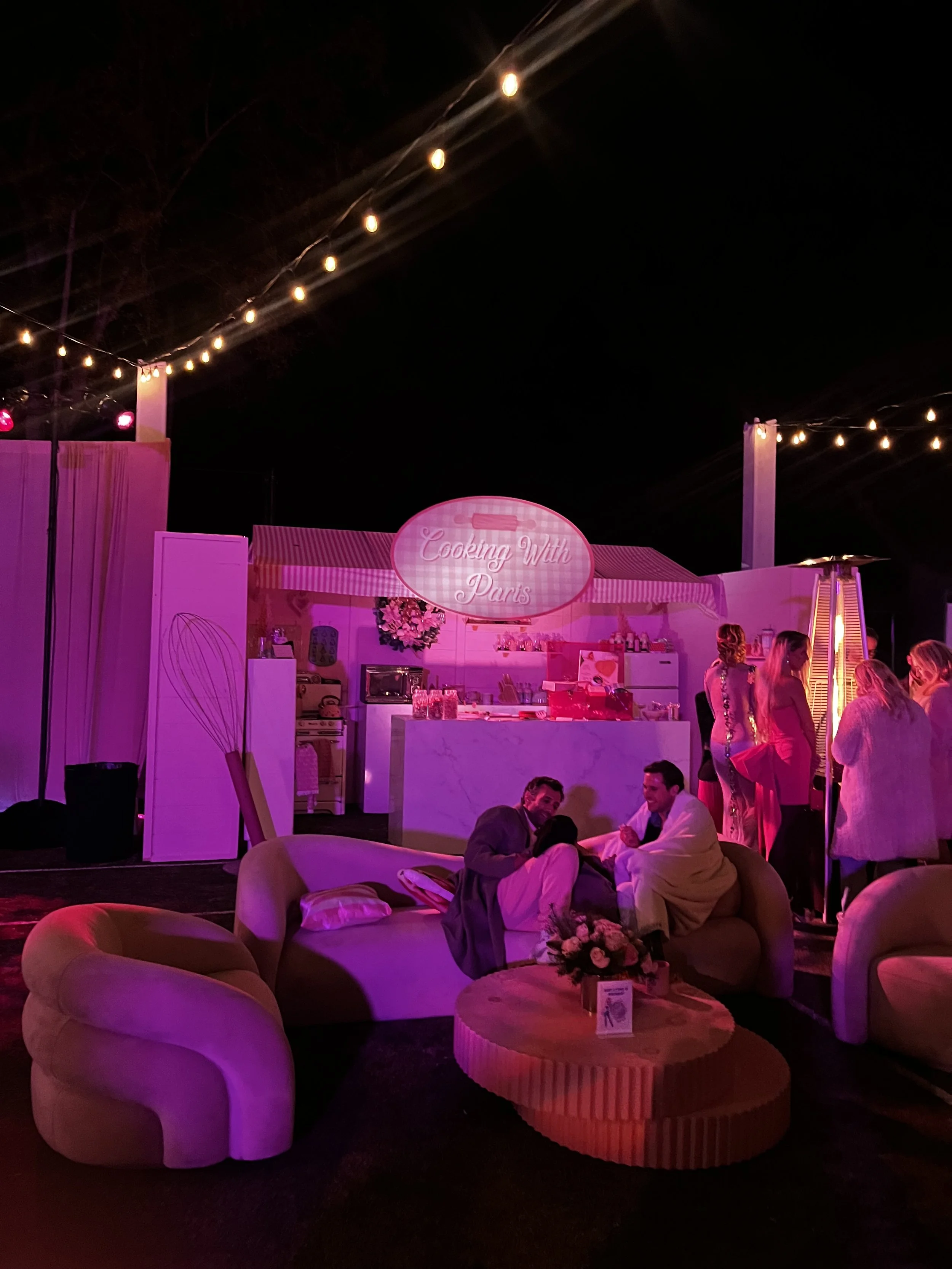 Nighttime outdoor event with pink lighting, a food stand labeled 'Cooking With Paris,' string lights, and people socializing on sofas and standing near a heater.