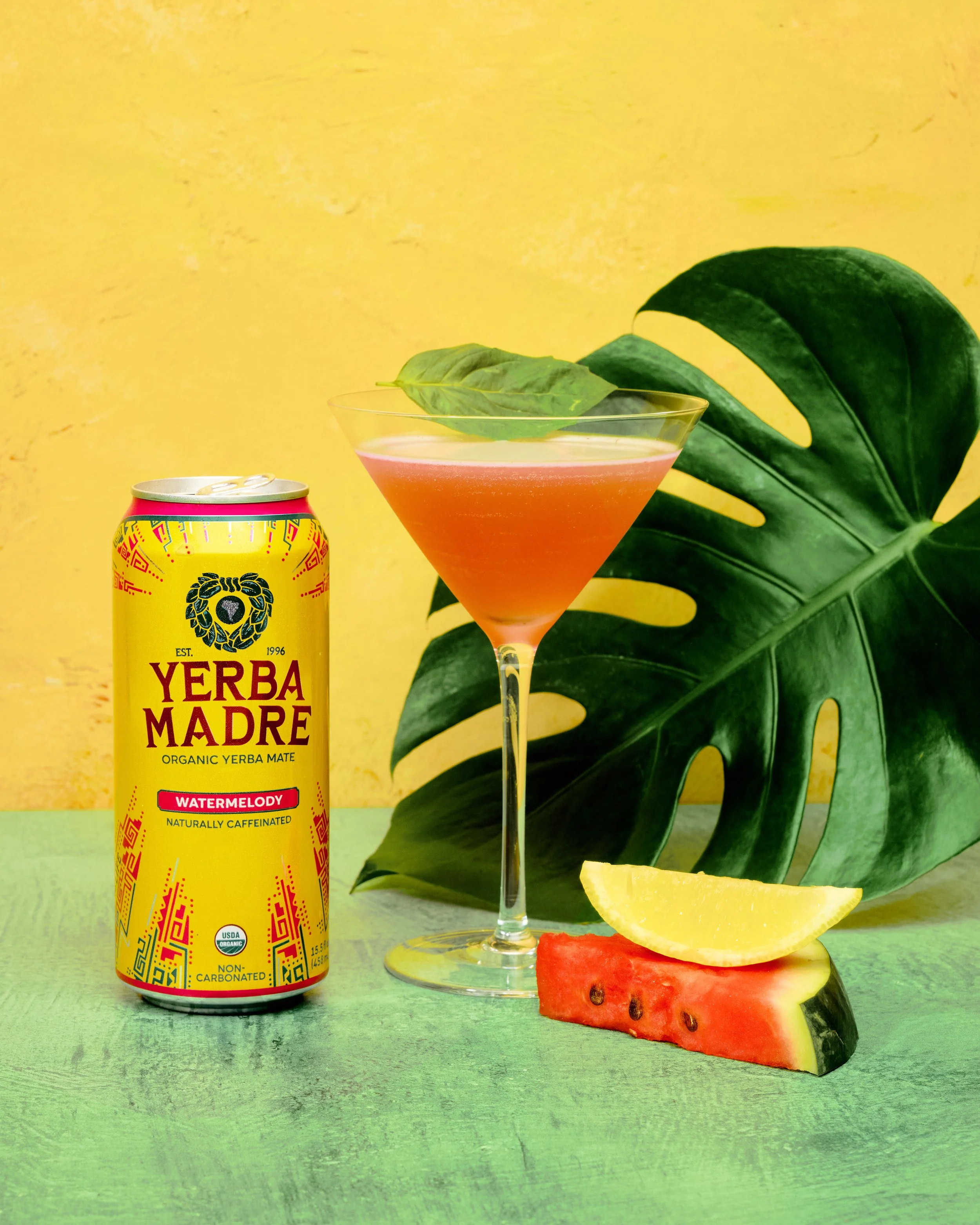 A yellow can of Yerba Madre Watermelon organic yerba mate, a pink cocktail garnished with a basil leaf, a large green monstera leaf, a slice of watermelon, and a lemon wedge on a green wooden surface with a yellow textured background.