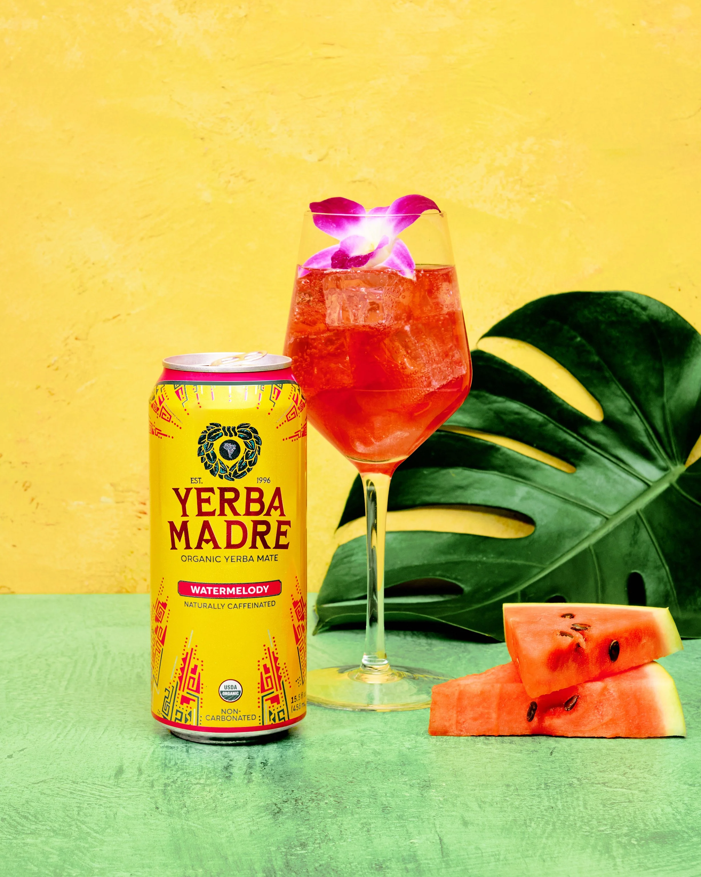 A can of Yerba Madre Watermelody flavor organic yerba mate next to a glass of watermelon mocktail with ice and purple flower garnish, with sliced watermelon and large green monstera leaf in the background on a green surface and yellow wall.