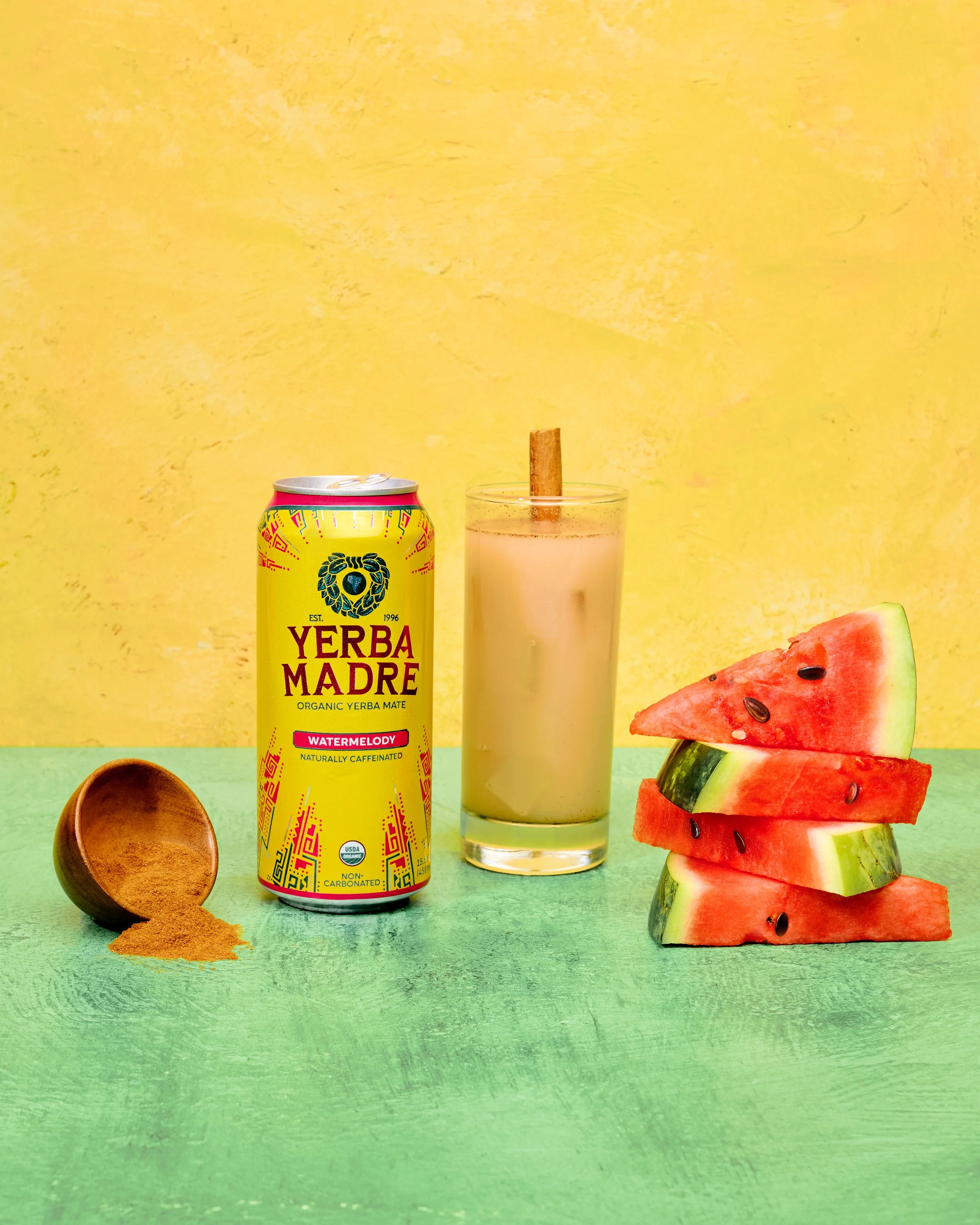 A can of Yerba Madre Watermelon flavored organic yerba mate, a glass with a cinnamon stick and a creamy beverage, and a stack of sliced watermelons with black seeds on a green surface against a yellow background.