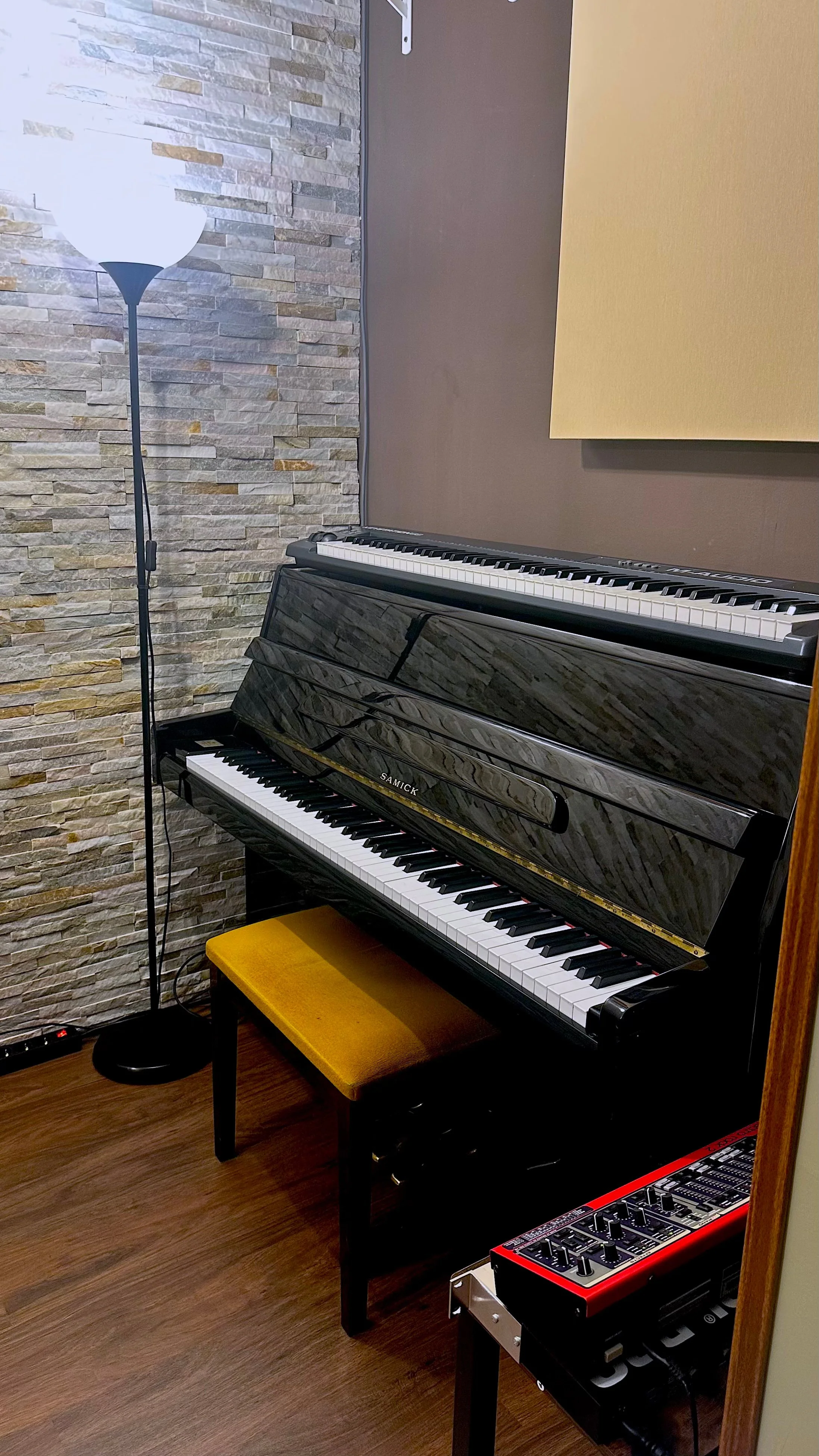 Un piano vertical negro sobre un banquillo amarillo en una sala de grabación. A la izquierda, una lámpara de pie y a la derecha, un teclado de efectos en una mesa.