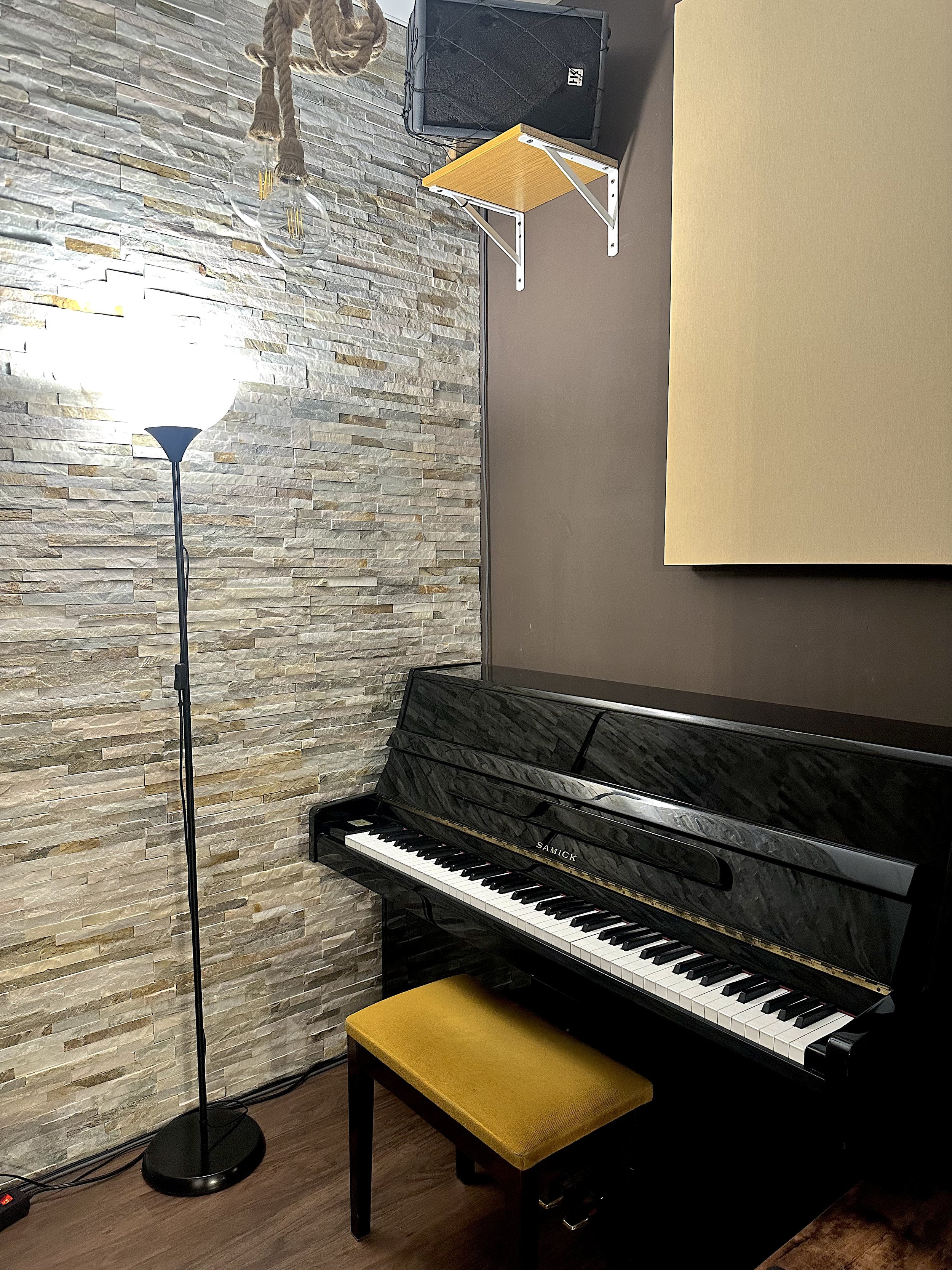 Un piano negro en una esquina de una habitación, con una banca amarilla delante, una lámpara de pie, y una pared de piedra decorativa. En la pared hay una pequeña mesa blanca y un altavoz.
