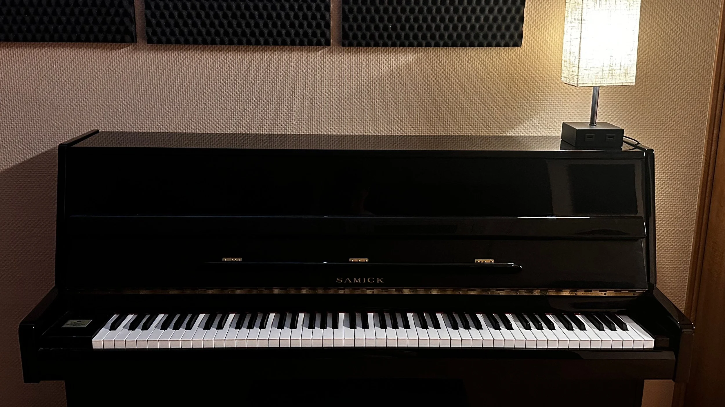 Piano negro con marca Samick, sobre fondo de pared beige con paneles acústicos y lámpara de mesa