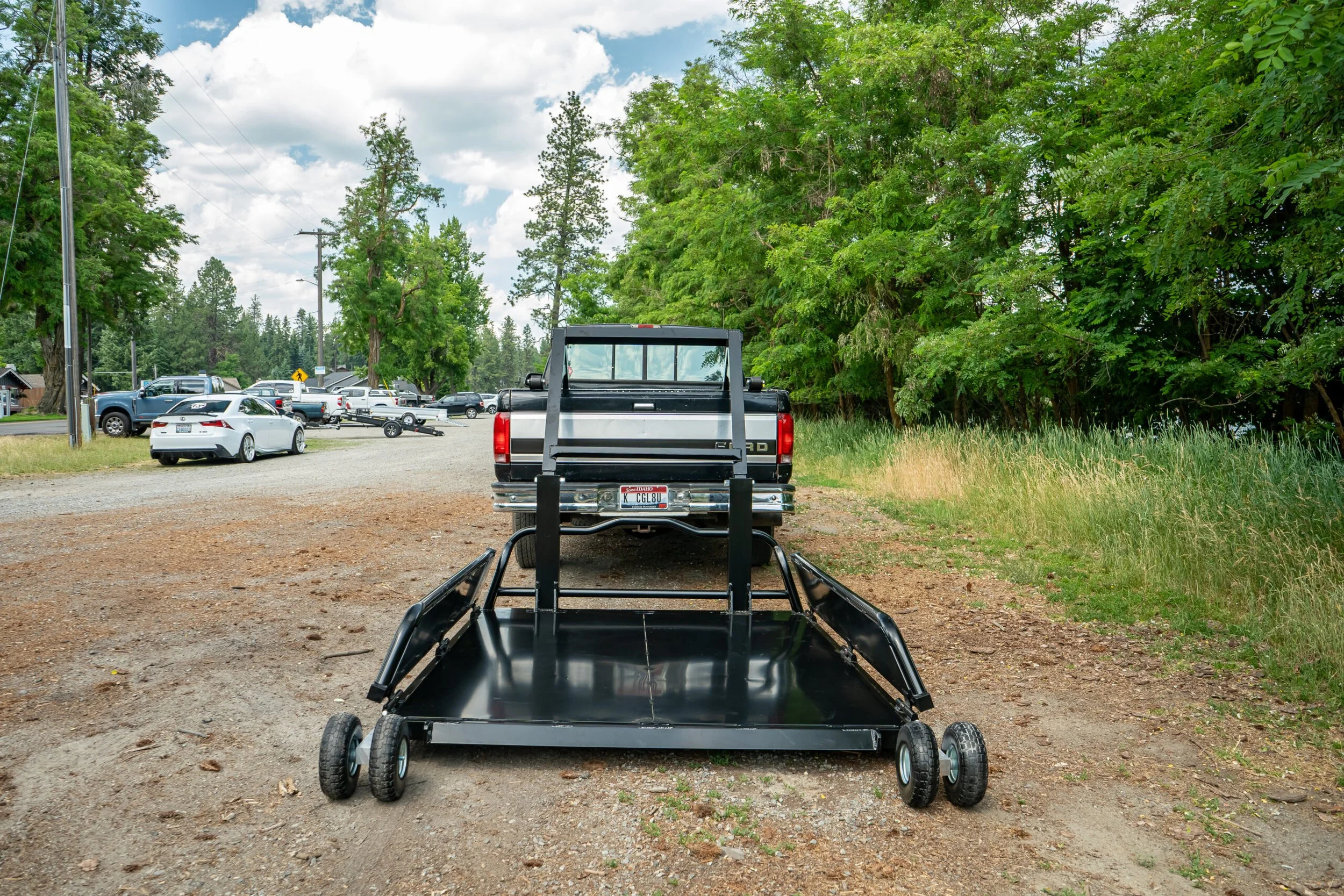 A black utility cart in a parking lot, with a pickup truck and several other vehicles and trees in the background.