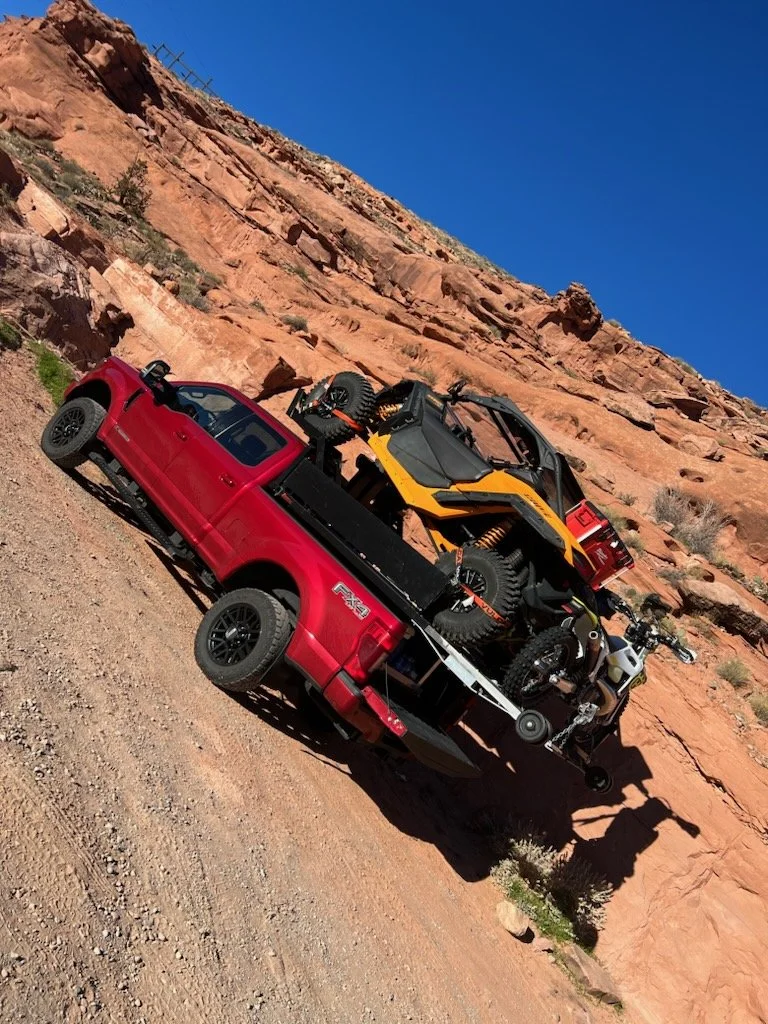 Red pickup truck with an ATV, a side-by-side utility vehicle, and a motorcycle loaded on its flatbed, parked on a dirt trail near red rock formations under a clear blue sky.