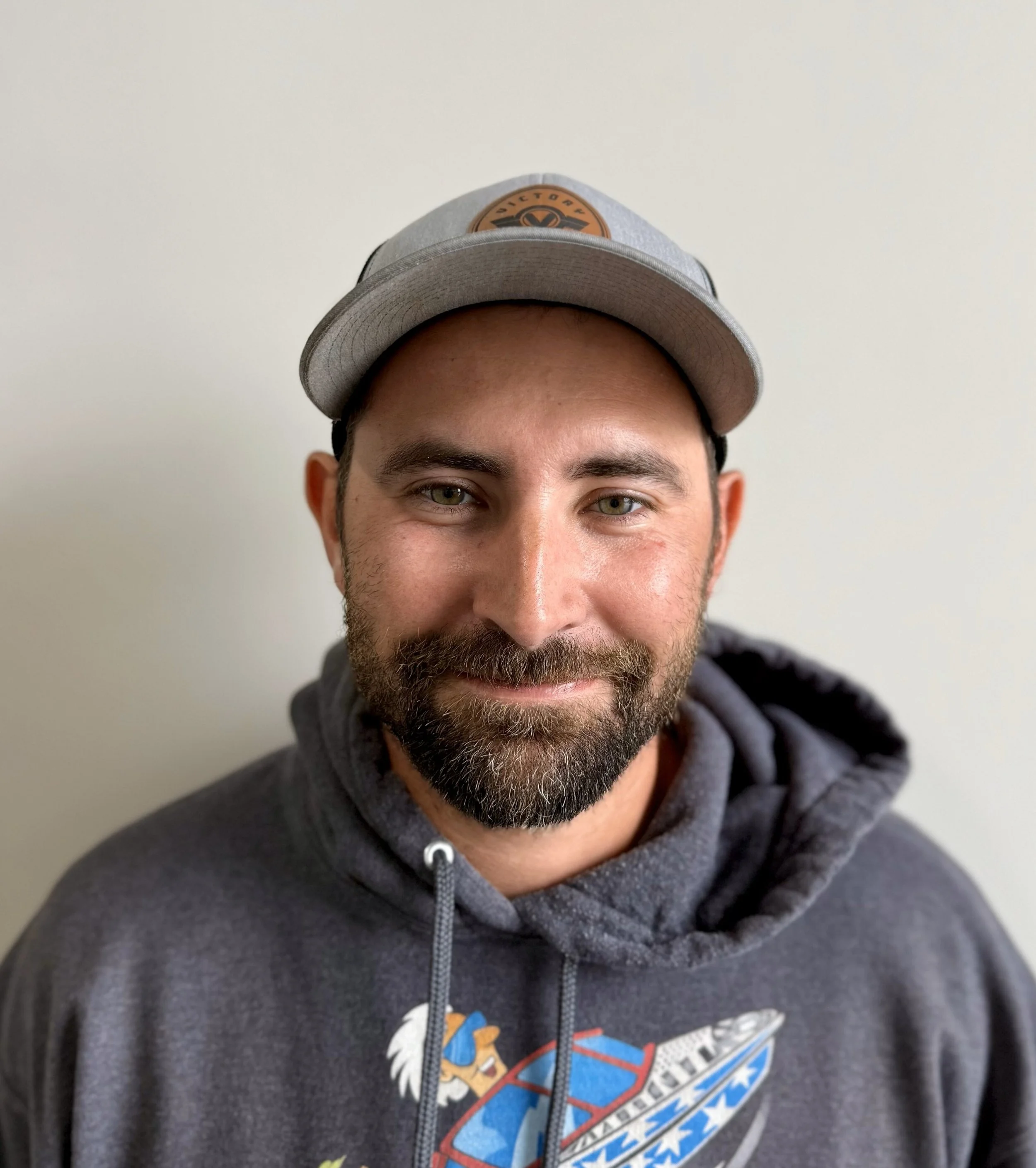 A man with a brown beard and mustache, wearing a gray baseball cap and a gray hoodie with a colorful graphic, smiles while looking at the camera, standing against a plain white wall.