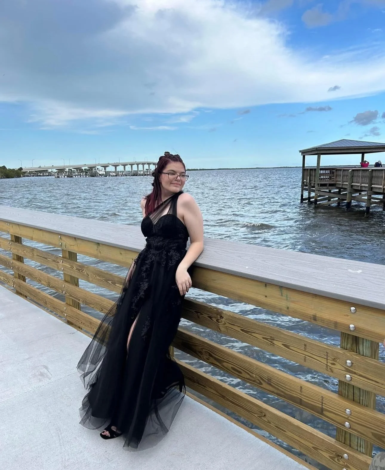 A young woman in a black dress with sheer details standing by a wooden railing on a waterfront pier. She is facing away from the water, with her eyes closed and a slight smile. The background features a large body of water, a bridge in the distance, 
