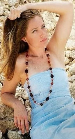 A woman with long blonde hair, sitting outdoors against a stone wall, wearing a light blue strapless dress and a beaded necklace, looking to the side with one arm raised and hand resting on her head.