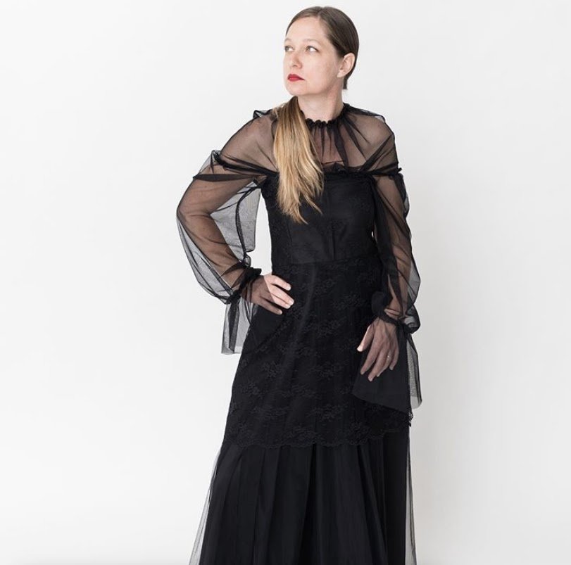 A woman with long blonde hair and red lipstick wearing a sheer black long-sleeved top and a layered black skirt standing against a plain white background.