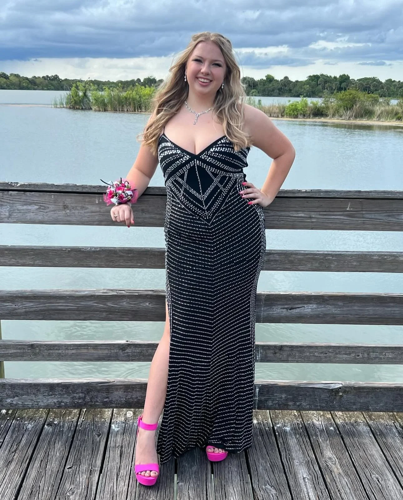 A young woman with long blonde hair wearing a sparkly black and white dress and pink high heels standing on a wooden dock by a body of water, posing with one hand on her hip and smiling.