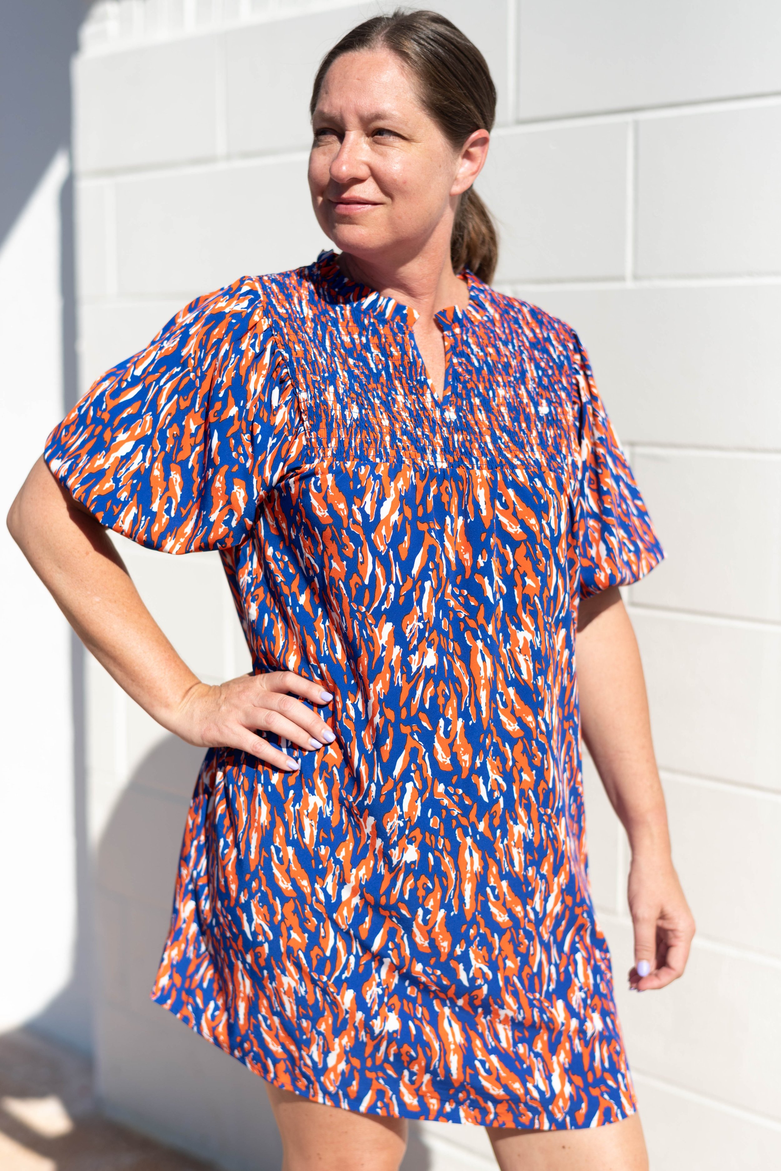 A woman in a colorful orange, blue, and white abstract patterned dress stands against a white wall, with one hand on her hip and looking to the side in Atlanta, GA