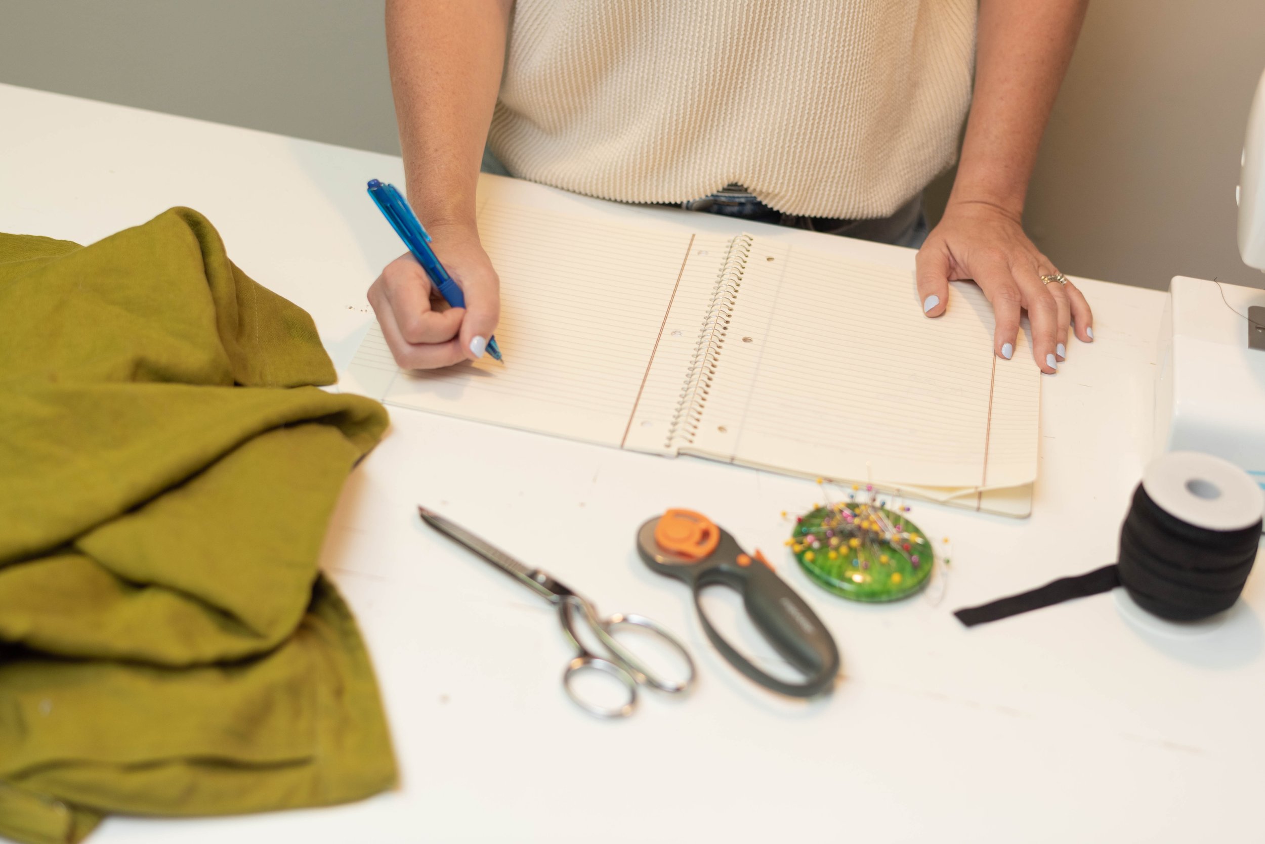 Person writing in a planner at a white table, surrounded by scissors, sewing pins, thread, and fabric.