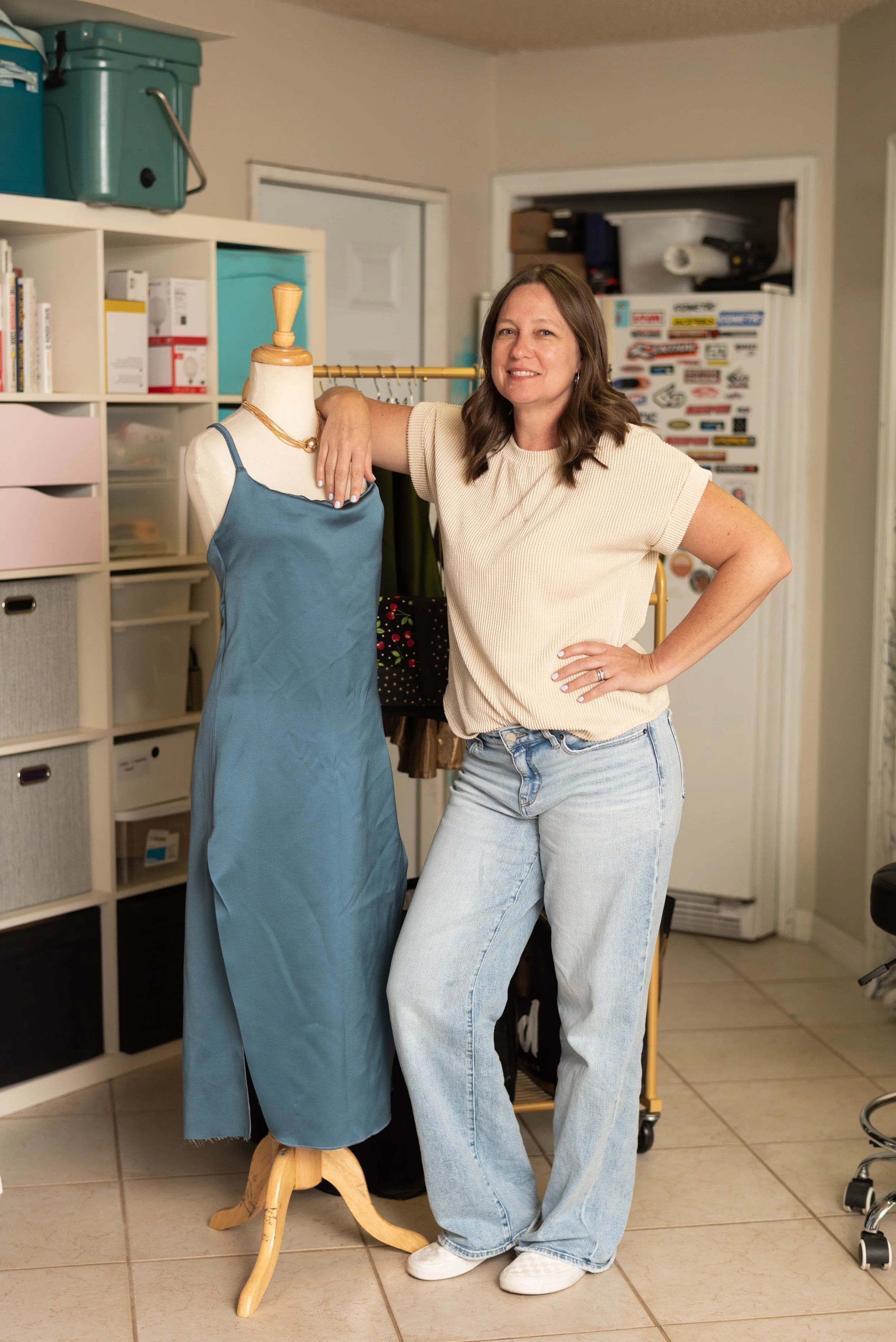 A woman with brown hair and a beige shirt standing next to a mannequin wearing a blue dress, in a room with shelves, boxes, and various items in the background in Atlanta, GA