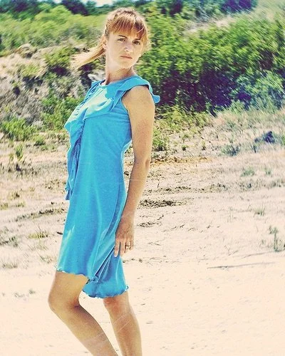 A woman in a bright blue dress standing on a sandy beach with green hills and shrubs in the background.