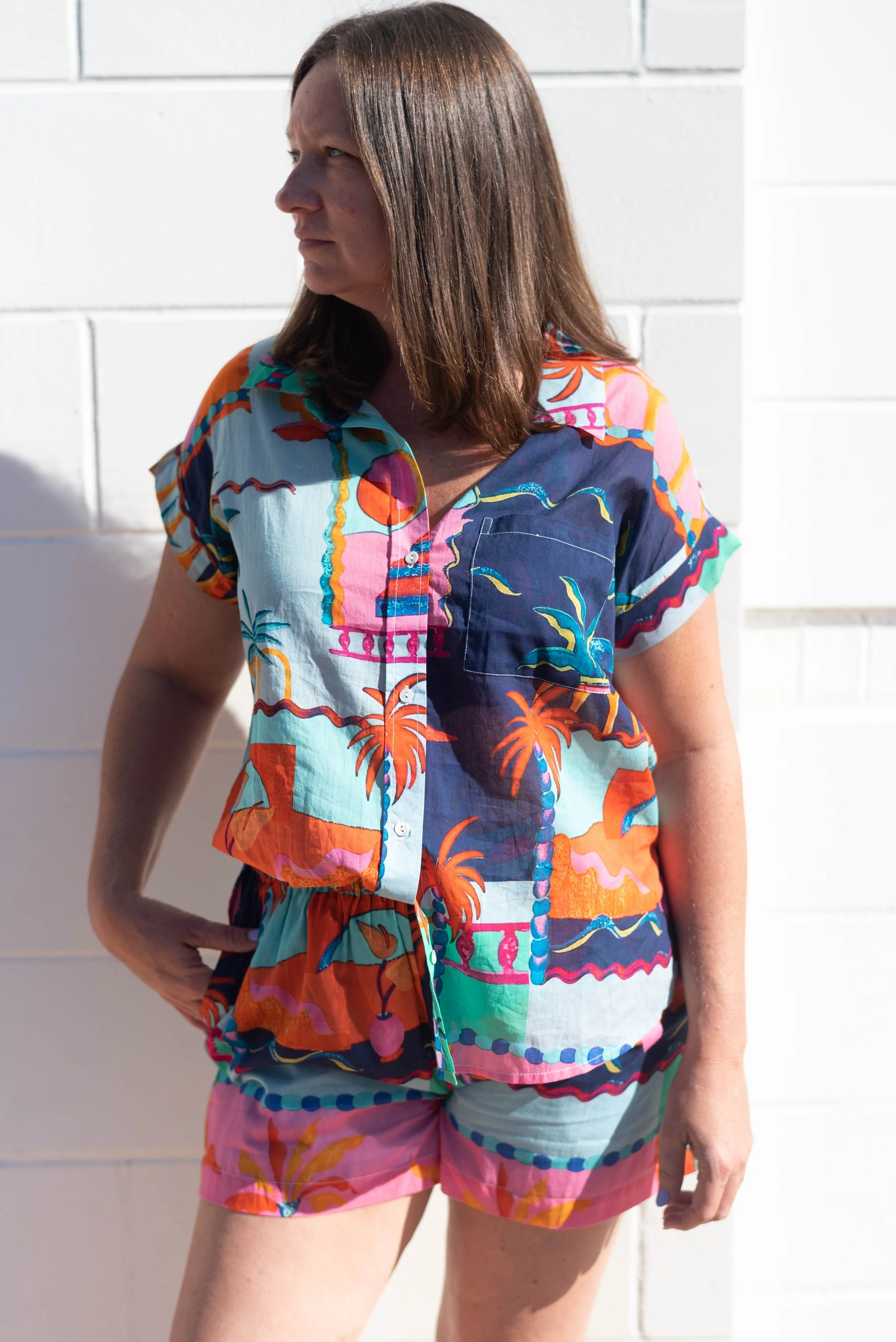 A woman wearing a colorful tropical print short-sleeve shirt and matching shorts, standing outdoors against a white brick wall in Atlanta, GA