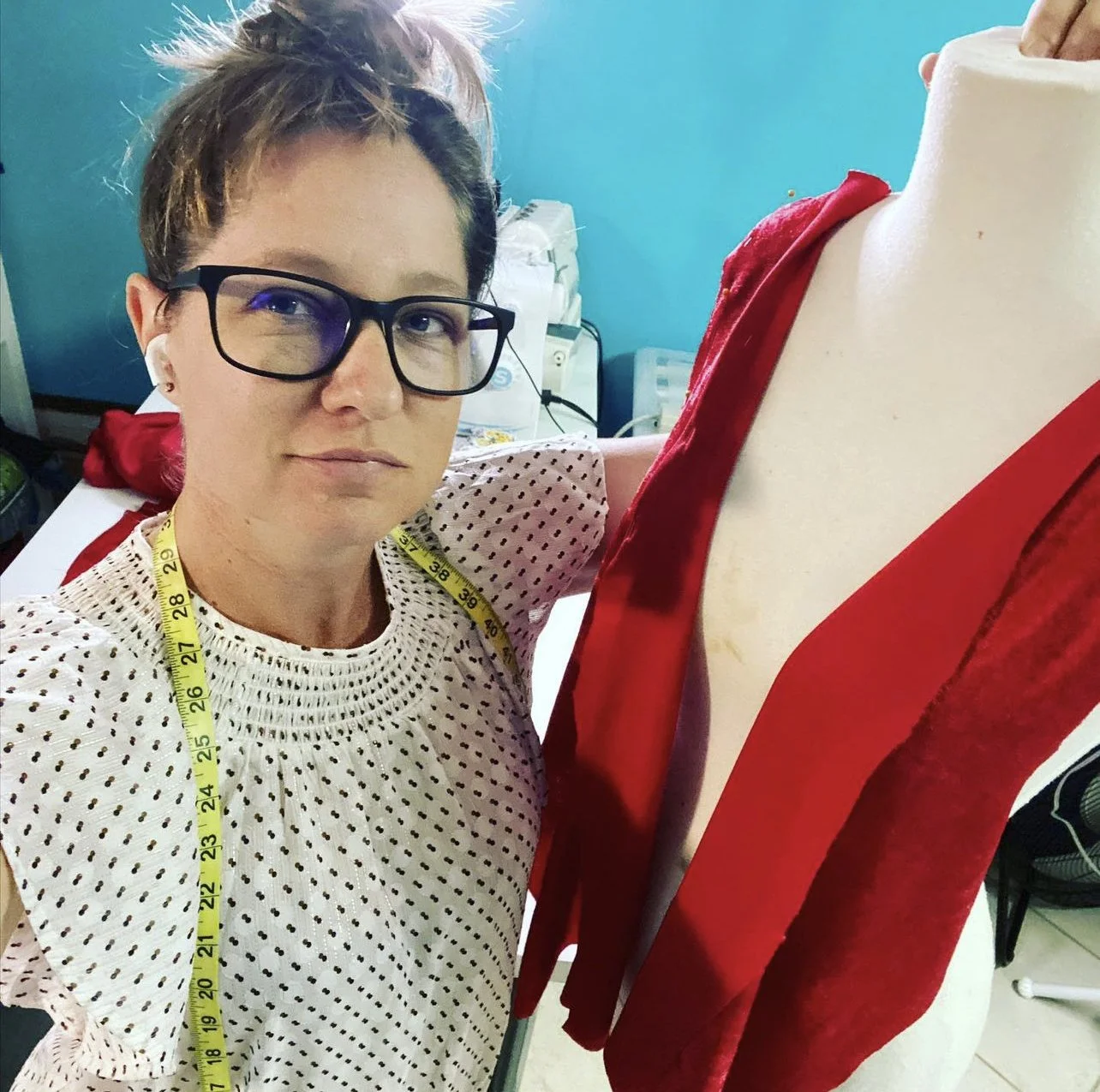 Woman with short hair, glasses, and white polka dot shirt sitting next to a dress form wearing a red garment, in what appears to be a sewing or fashion design workspace.