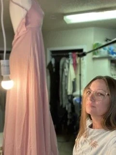 A woman with long hair and glasses holding a pink dress in a room with a closet and various items in the background.