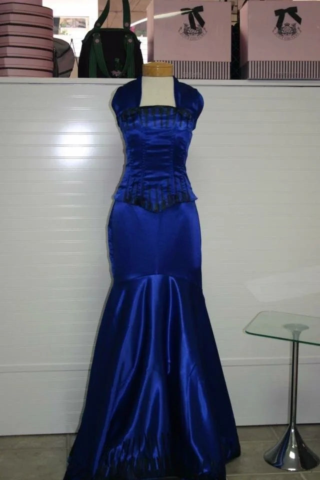 A blue satin evening gown with a fitted bodice and a mermaid-style skirt displayed on a mannequin. The bodice has a ruched design and a matching bolero jacket.