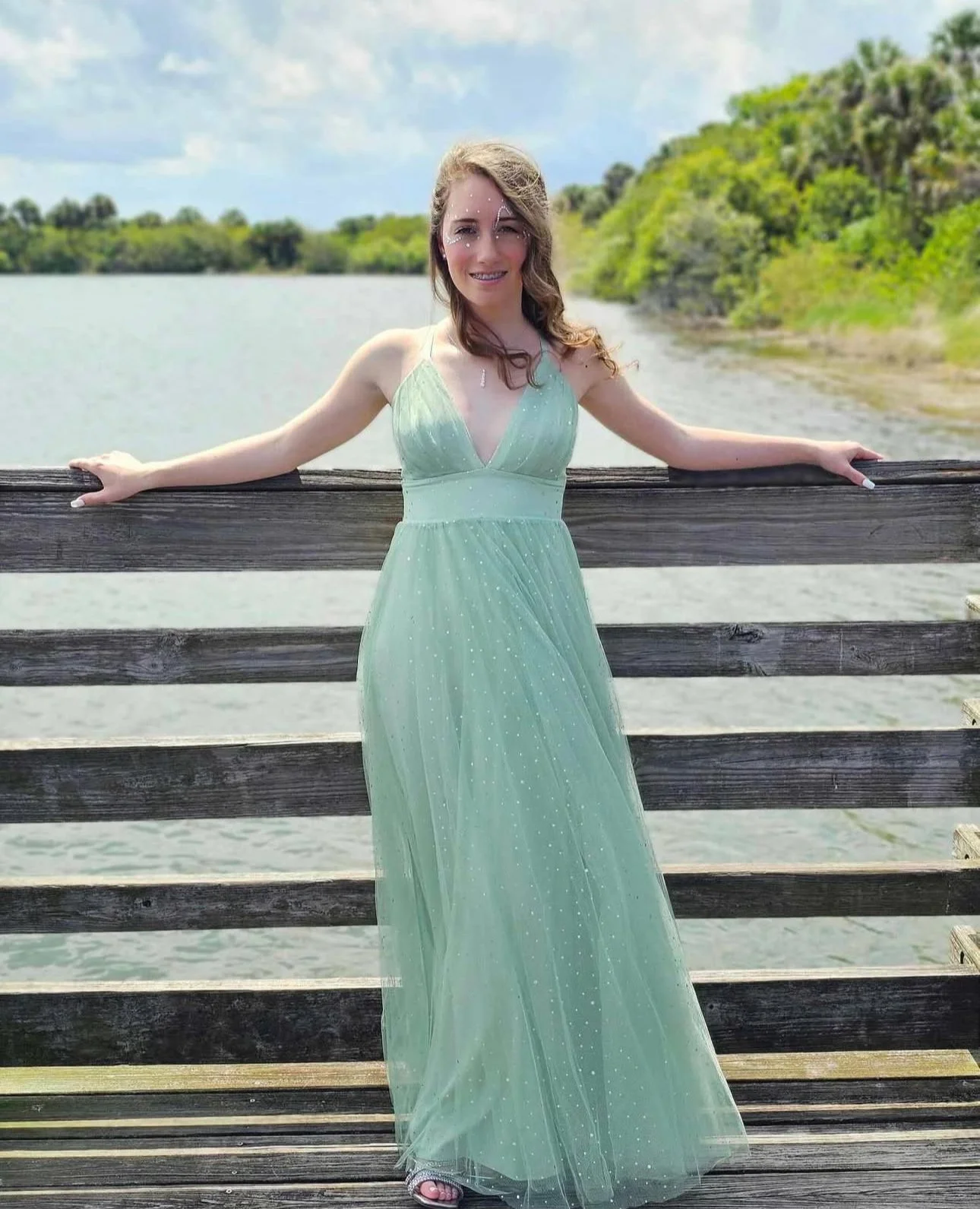 A young woman in a light green, flowy dress standing on a wooden dock by a body of water with green trees in the background.