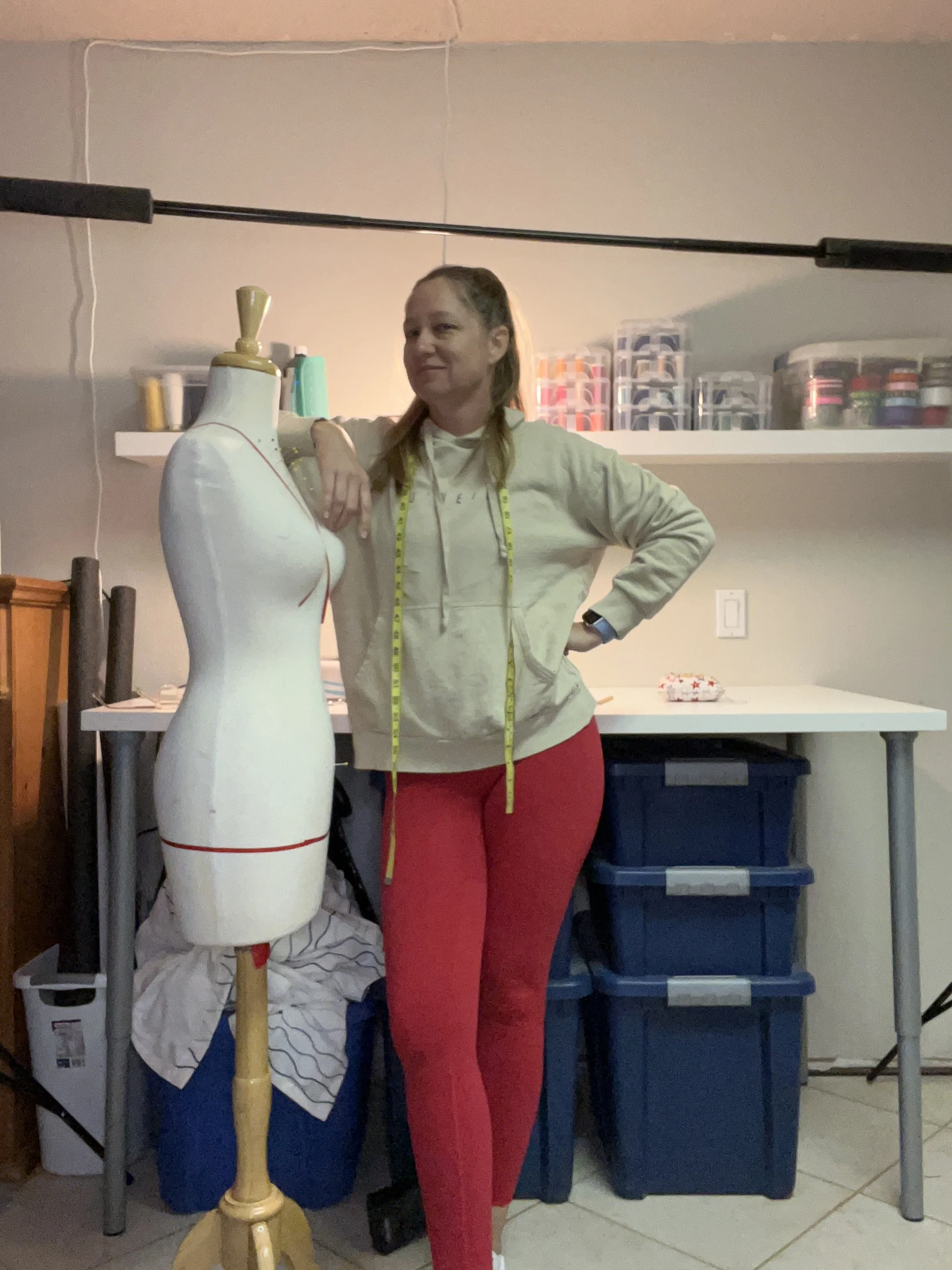 A woman stands next to a dressmaker's mannequin with a measuring tape draped around her neck, in a room with shelves and plastic storage bins.