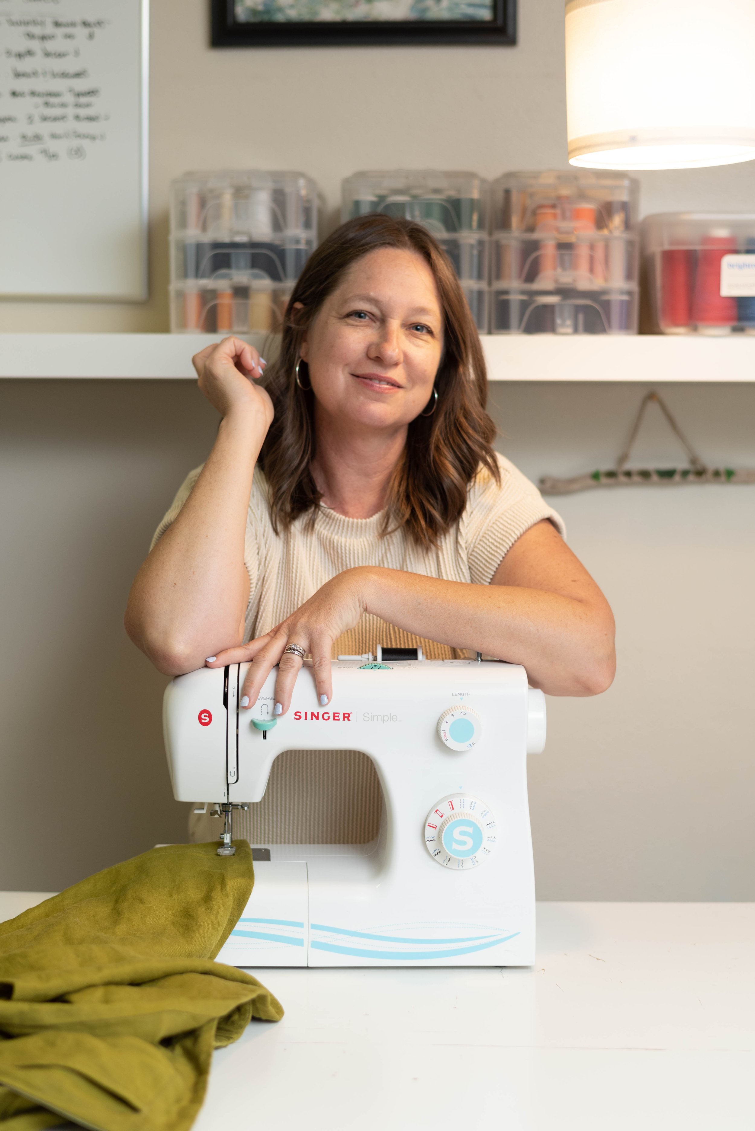 A woman with brown hair and hoop earrings smiling at the camera, resting her chin on her hand, sitting behind a white sewing machine on a table, with a piece of yellow fabric in front of her, in a room with storage bins and a whiteboard in the background.