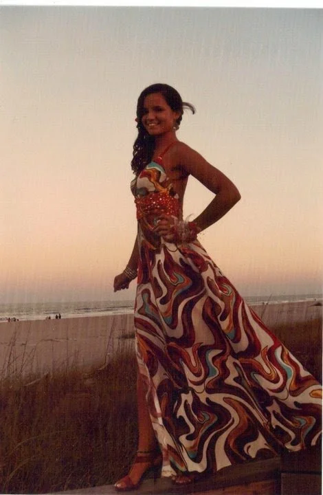 A woman in a colorful, patterned maxi dress standing on a beach at sunset, smiling and posing with her hand on her hip.