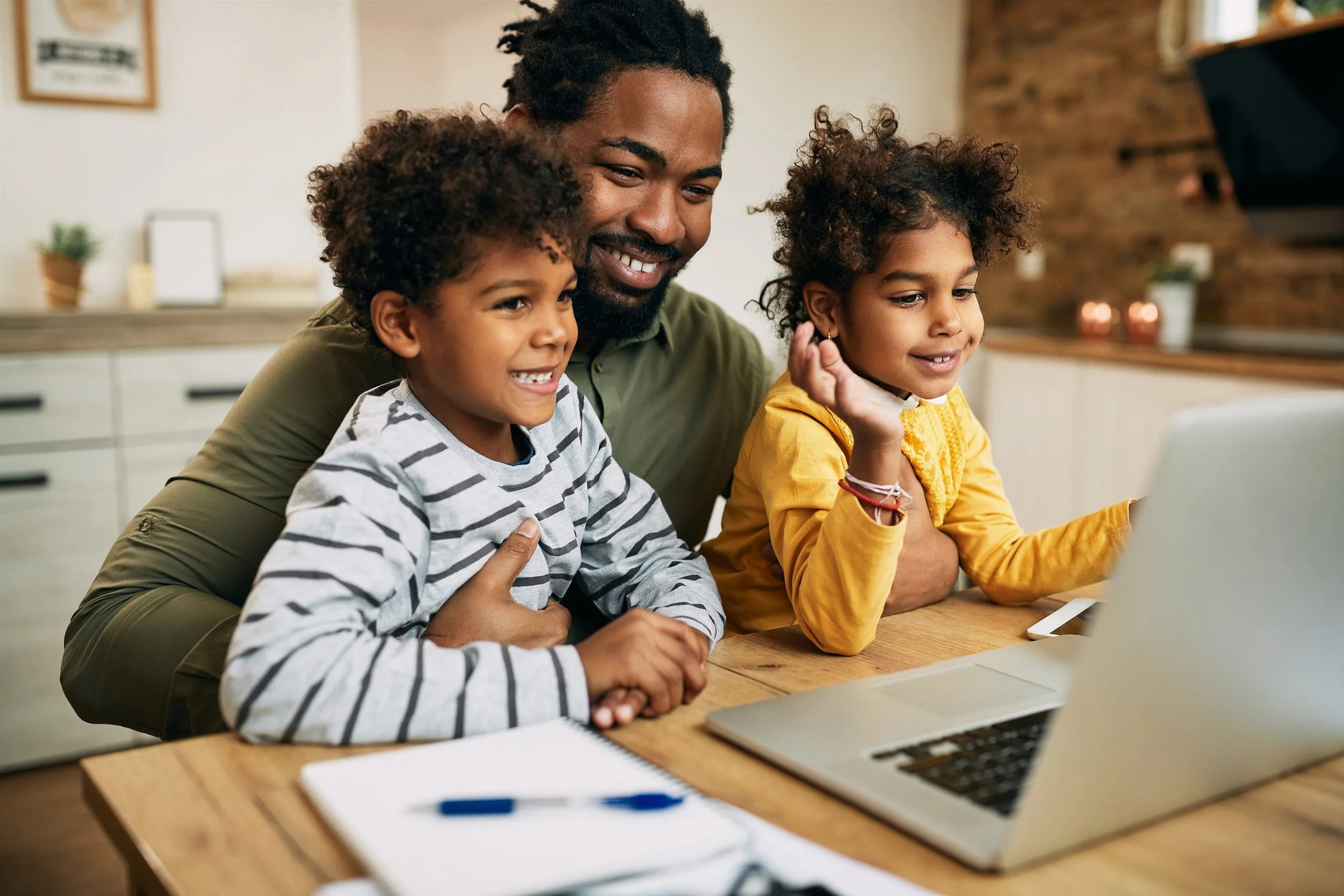 A man smiling with two children looking at a laptop screen in a cozy kitchen.