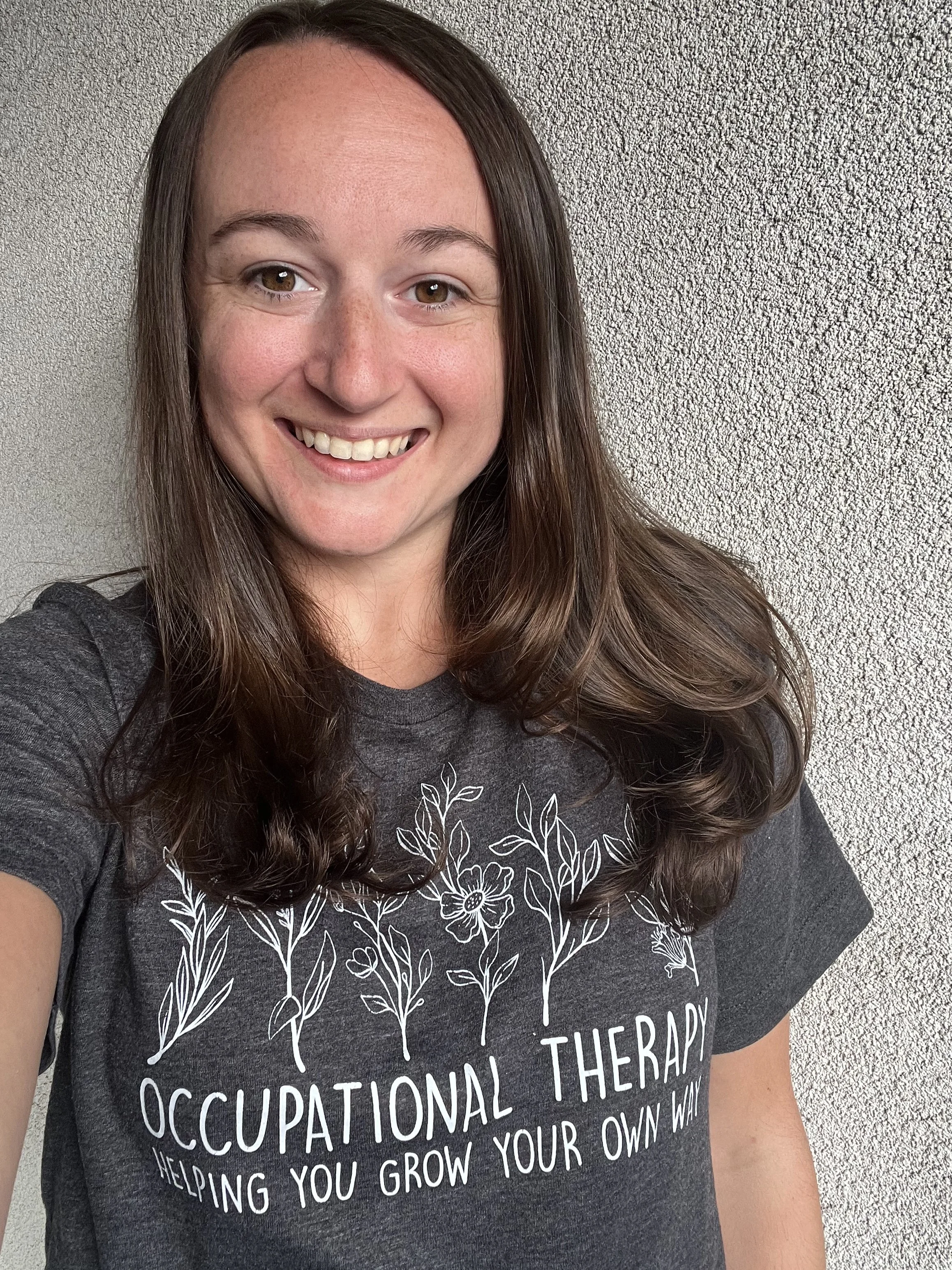 A woman with brown hair taking a selfie against a textured gray wall, wearing a dark gray T-shirt with white botanical print and text related to occupational therapy.