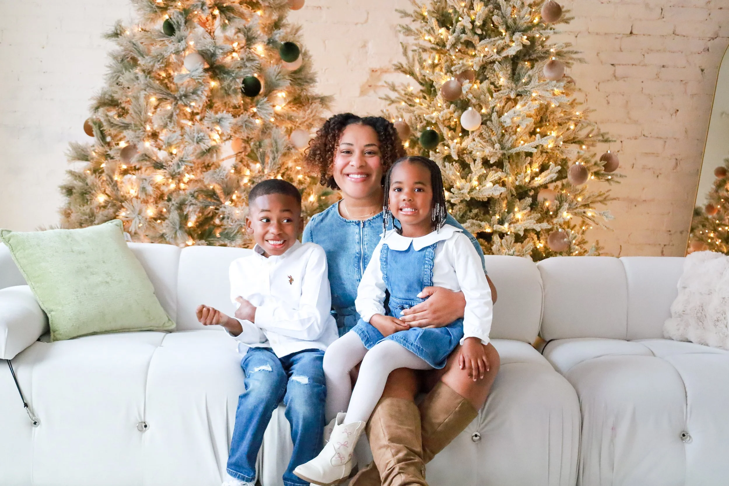 A woman with curly hair and two children sitting on a white couch in front of Christmas trees decorated with ornaments and lights.