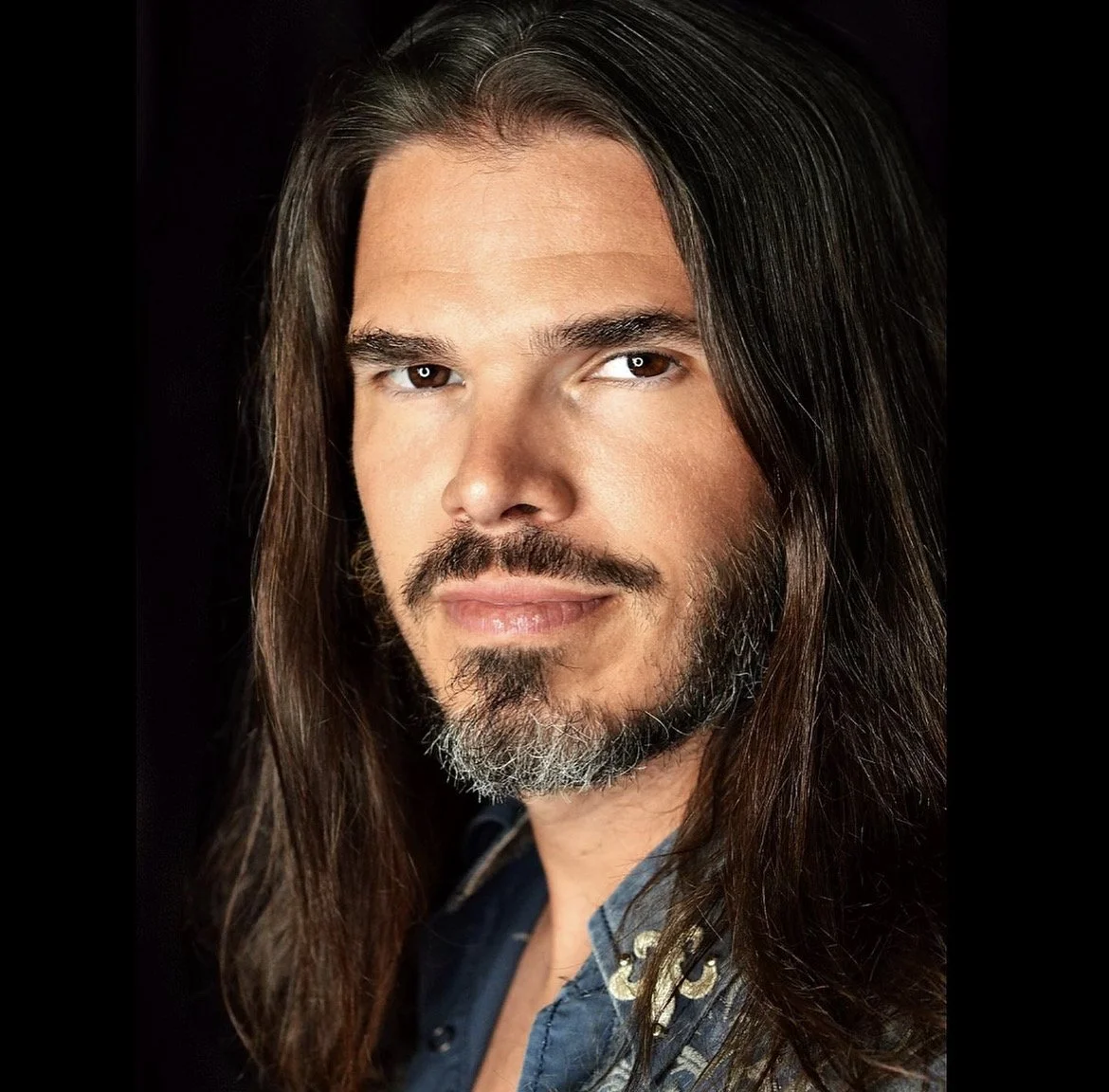 A man with long dark hair, a beard, and mustache, wearing a blue shirt with a floral pattern, looking confidently at the camera.