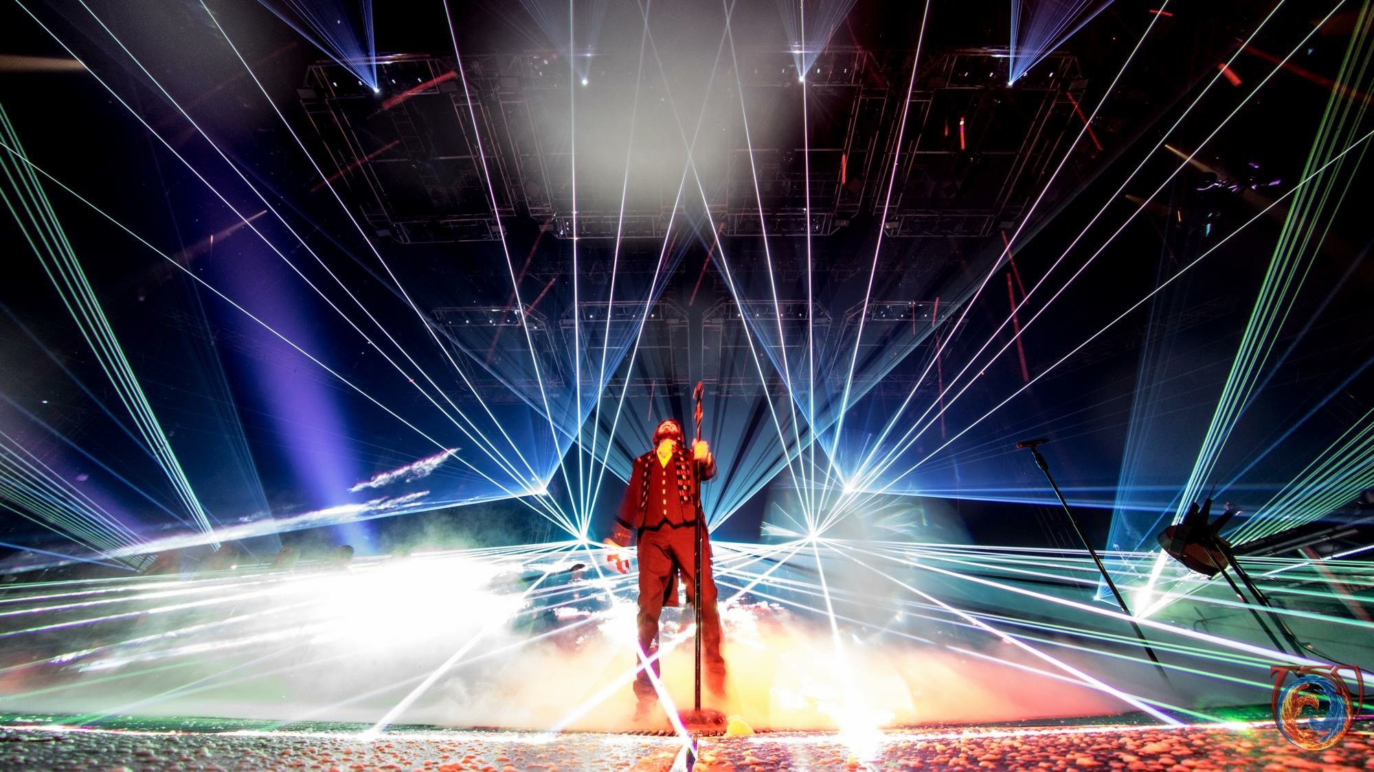 Performer on stage with a staff, surrounded by colorful laser lights and stage smoke.