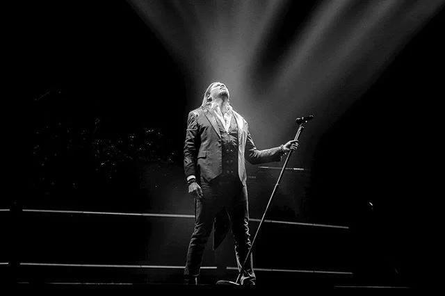 A singer in a tuxedo holding a microphone stand on stage with dramatic lighting.