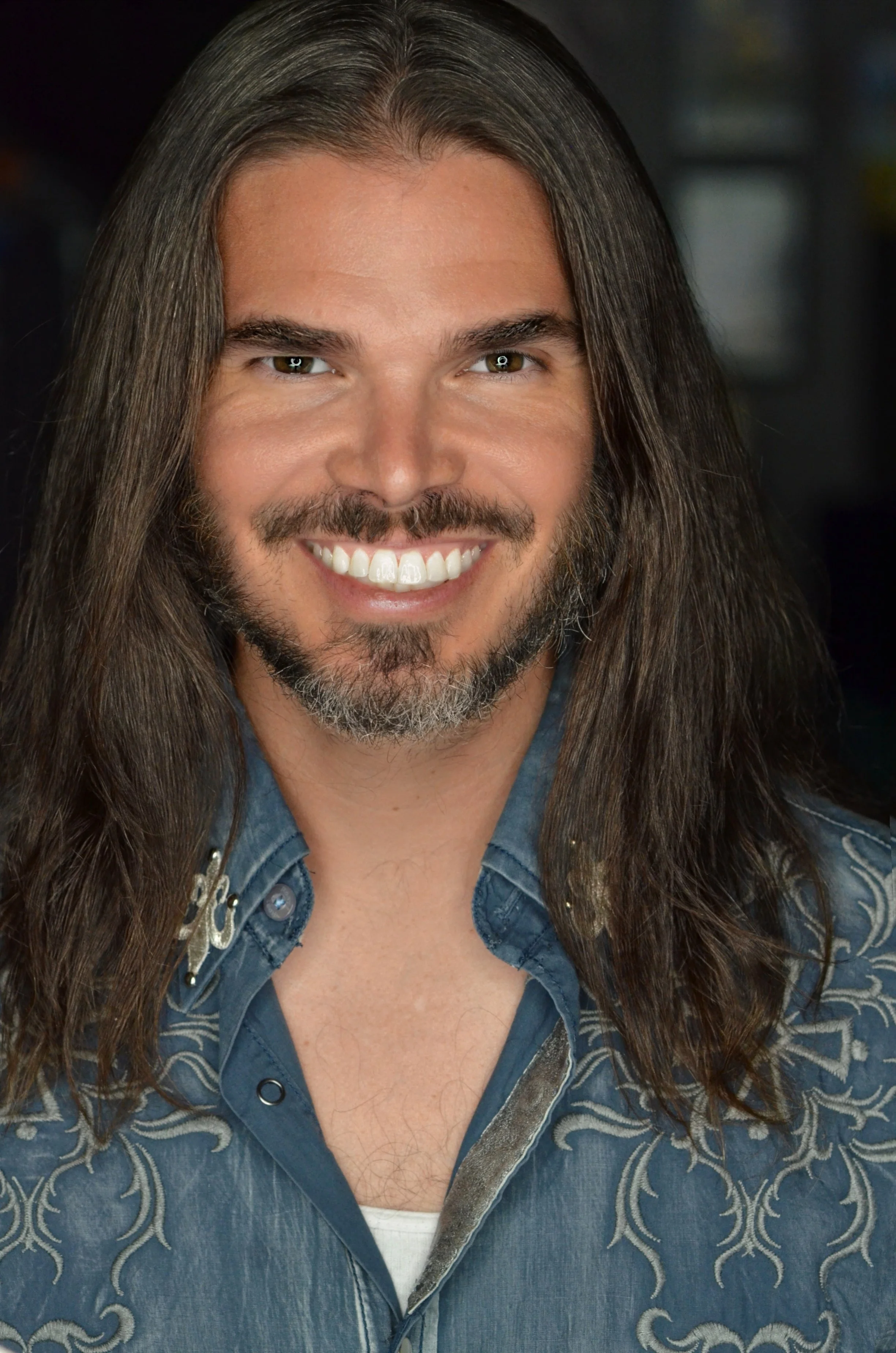 A man with long brown hair, a beard, and a bright smile, wearing a denim shirt with embroidery detail, against a blurred dark background.