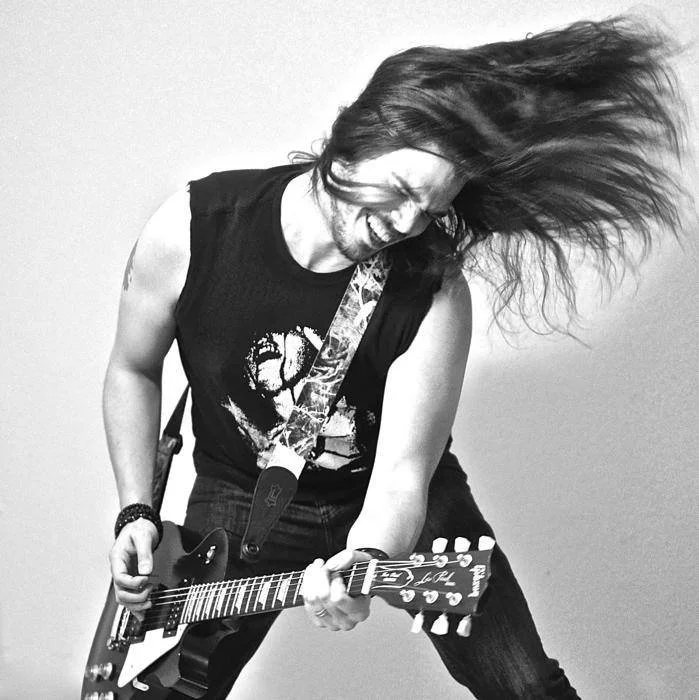 Person playing an electric guitar, head tilted with hair flying, wearing a sleeveless shirt and a wristband.