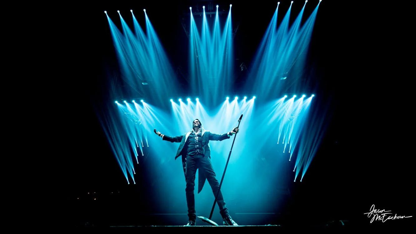 Man in a suit holding a microphone stand on stage with blue spotlights overhead
