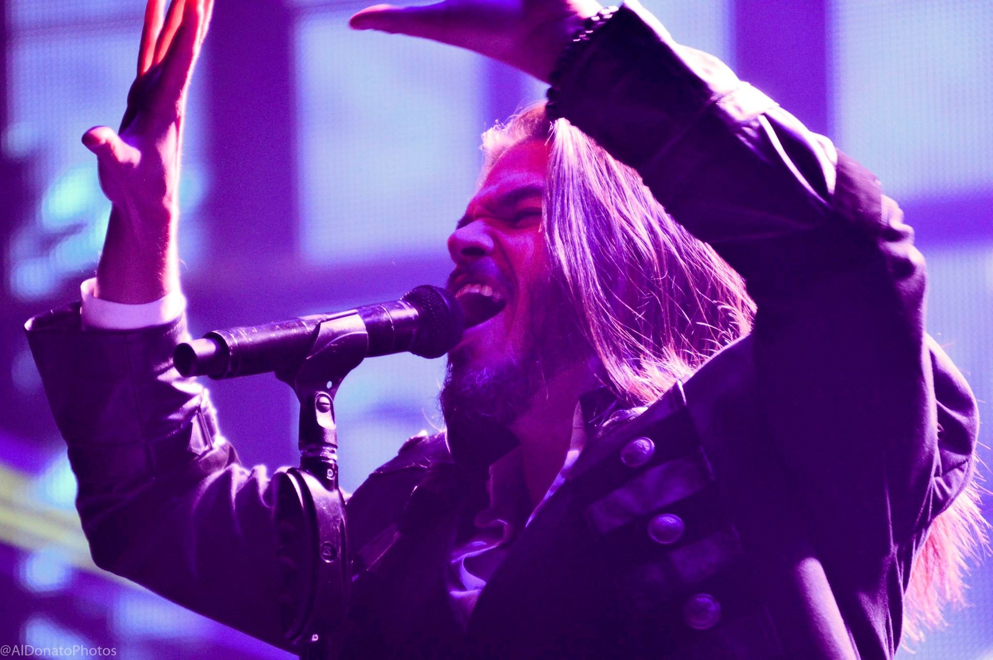 Male singer with long hair passionately singing into a microphone, wearing a black leather jacket with purple stage lighting.
