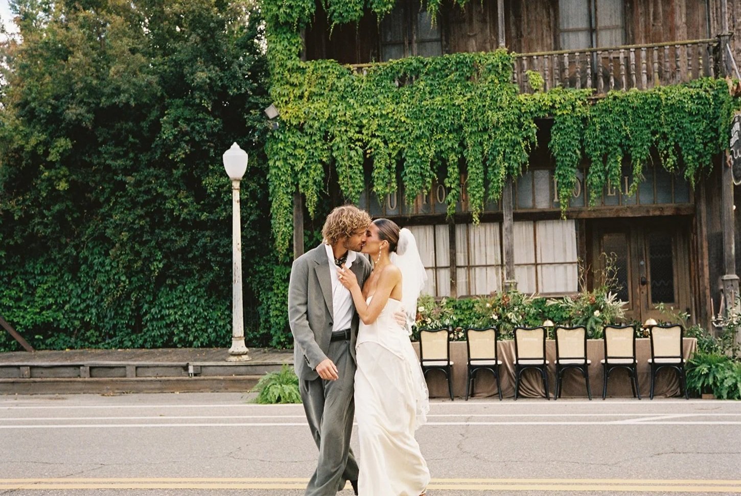 A couple dressed in wedding attire sharing a kiss in the middle of a street, with an old building covered in green ivy and an outdoor table with chairs in the background.