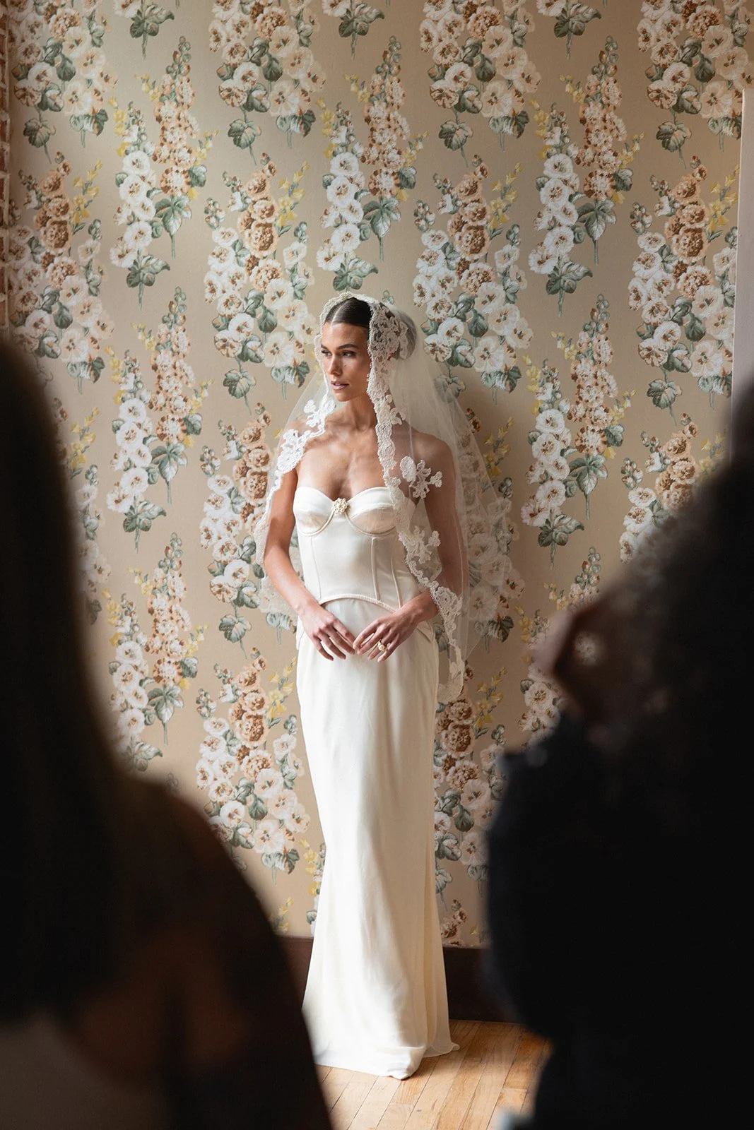 Bride in a wedding dress with a veil standing against a floral patterned wallpaper, with two blurred figures in the foreground in bridal suite at 1880 Union Hotel a historic wedding venue in Southern California.