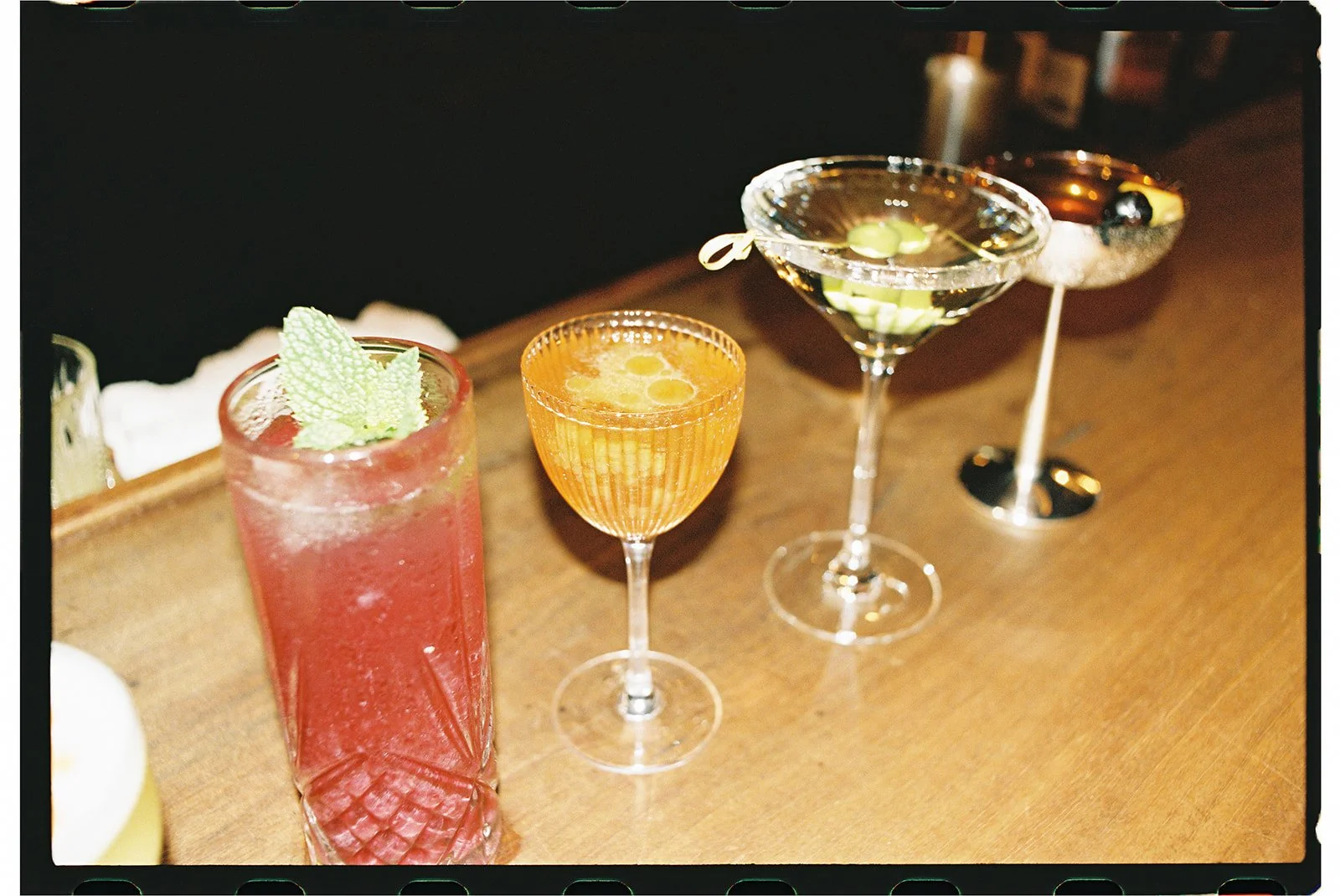 Four colorful cocktails on a wooden bar counter, including a pink drink with mint garnish, an orange drink, a martini with olives, and a dark-colored cocktail with a cherry and slice of orange.