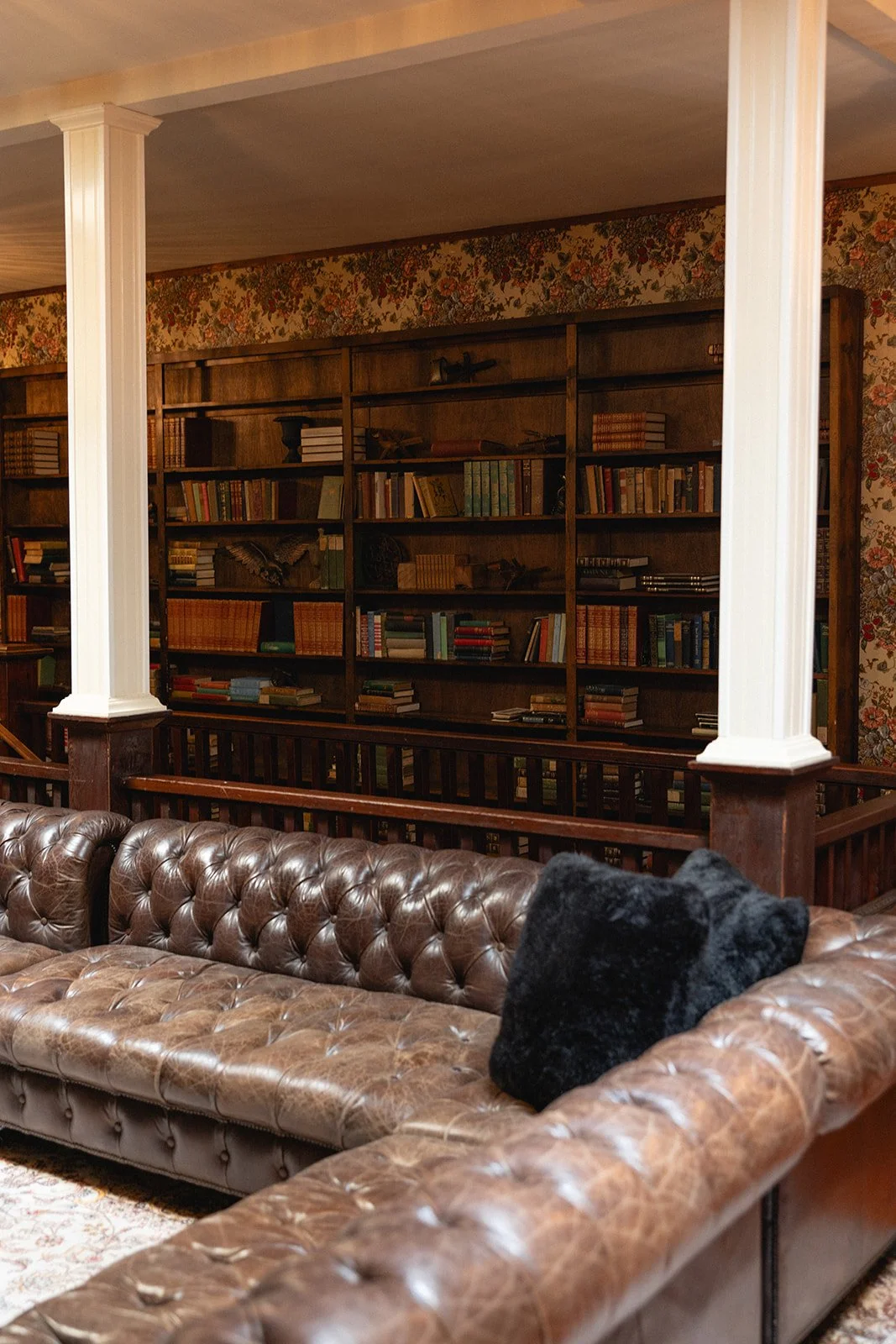 A vintage lounge with a tufted leather sofa, a black pillow, wooden bookshelf filled with books, floral wallpaper, and white columns at 1880 Union Hotel in Santa Barbara Wine Country.