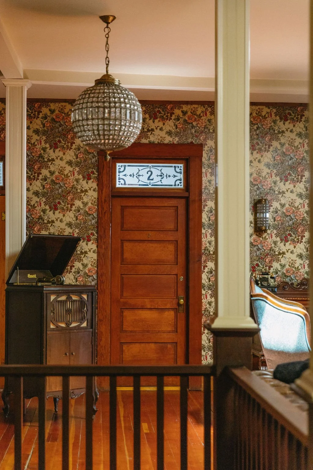 Interior view of a vintage hotel with floral wallpaper, a wooden door with a decorative glass window, a hanging chandelier, a record player on a wooden stand, and a blue armchair at 1880  Union Hotel a wedding venue outside Los Angeles California