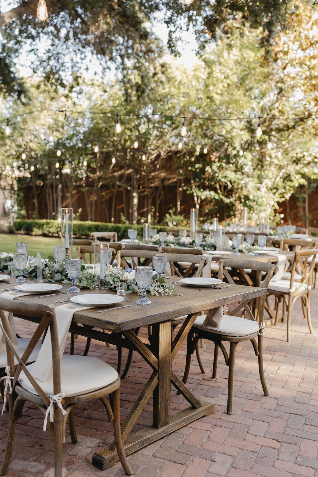 Outdoor wedding reception with decorated tables, chairs, and candle centerpieces under string lights and large trees on a brick patio at 1880 Union Hotel a premier all-inclusive wedding venue with lodging in Santa Barbara.