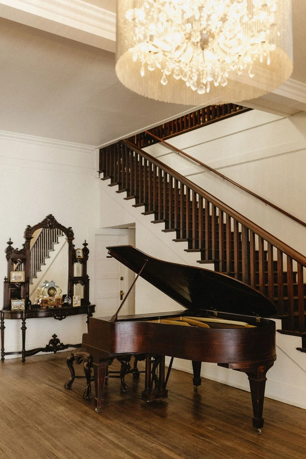 A grand piano with an open lid in a room with wooden floors, a staircase with a wooden railing, and an ornate mirror with a shelf holding framed photos and decorative items, illuminated by a chandelier at 1880 Union Hotel wedding venue with lodging.