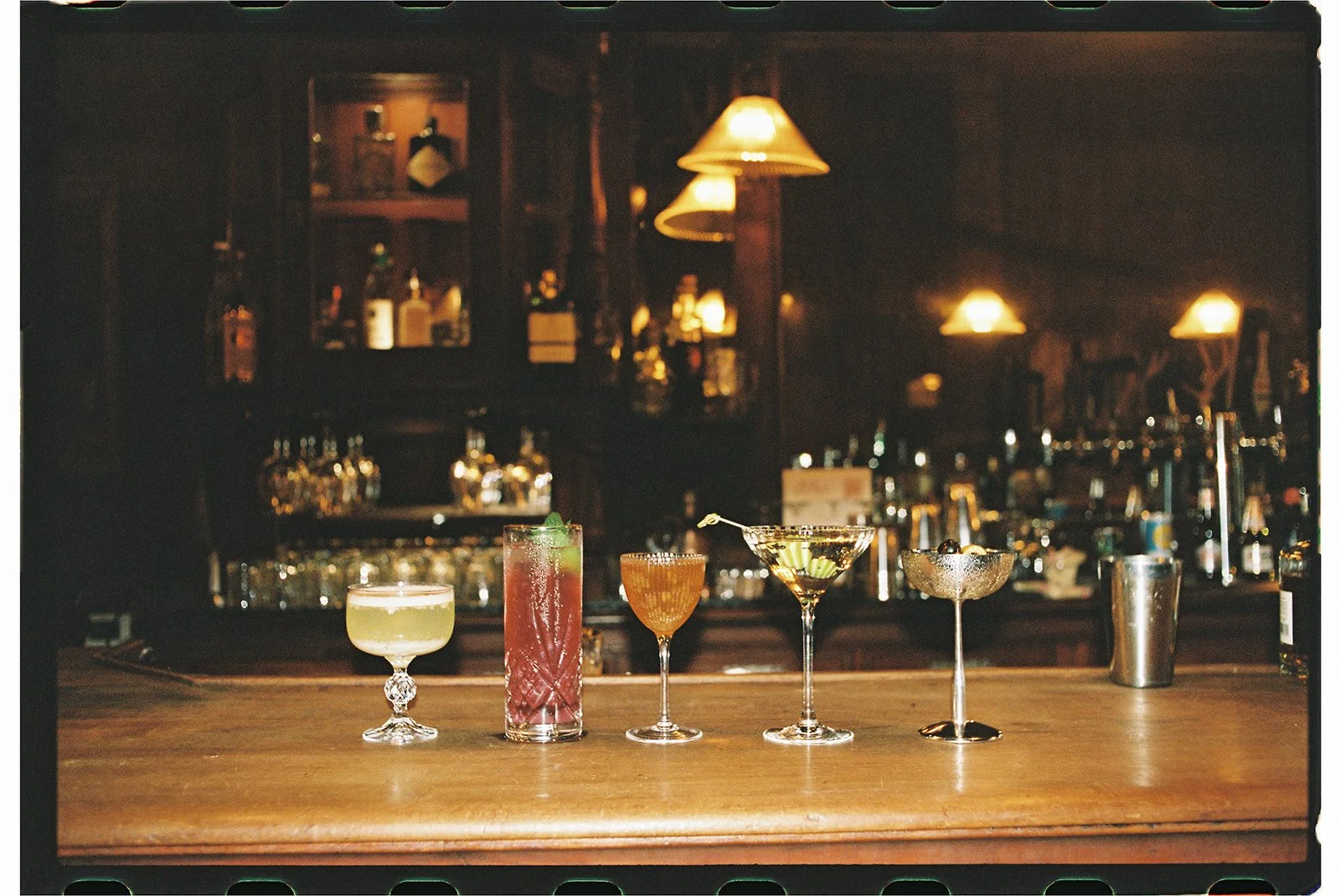 Seven colorful cocktails lined up on a wooden bar counter in a dimly lit bar with warm lighting and liquor bottles in the background at the Saloon in 1880 Union Hotel in Los Alamos, California.