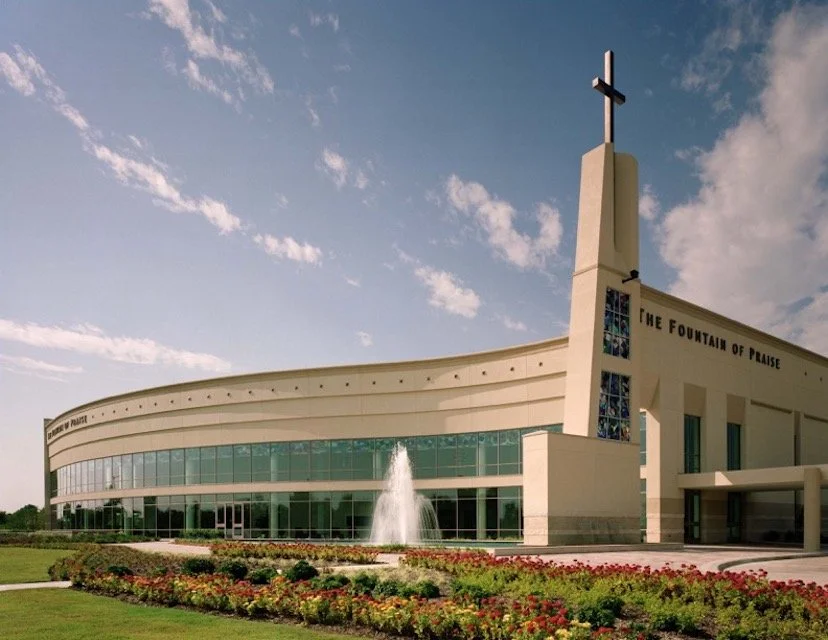 Exterior view of The Fountain of Praise church building with a cross on top, large stained glass windows, and a water fountain in front, surrounded by a flower bed under a partly cloudy sky.