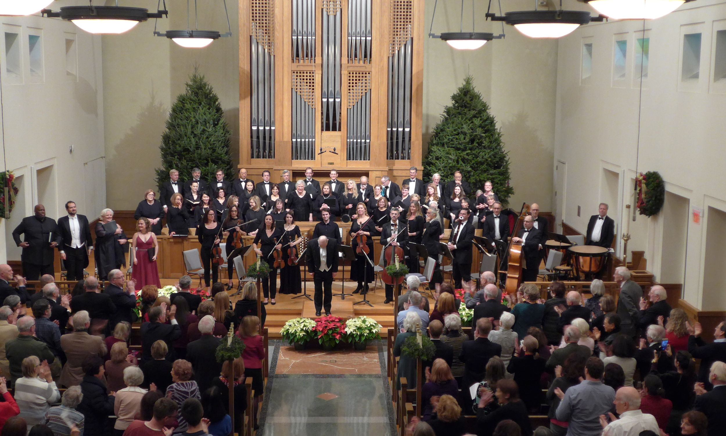 A large choir and orchestra on stage in a church decorated for Christmas, with Christmas trees and wreaths, after a performance, with audience members applauding.