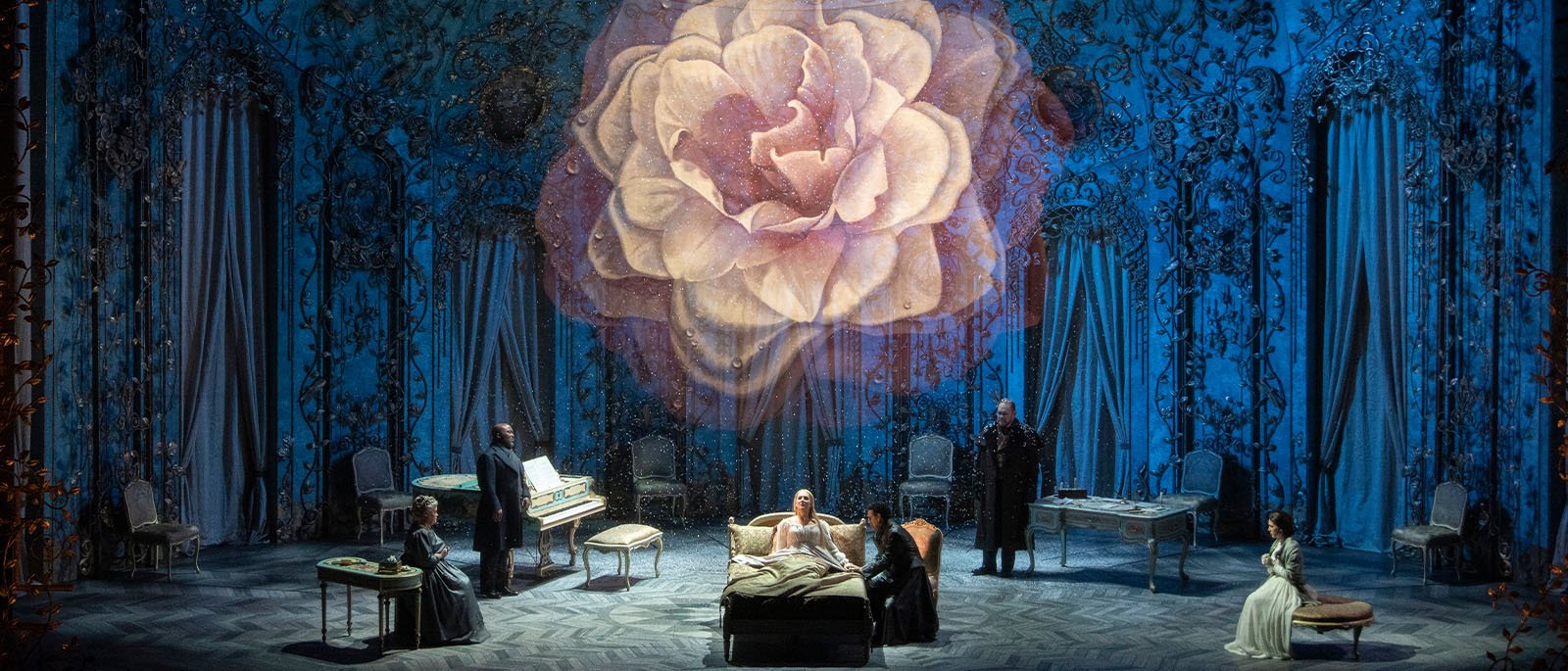 A theatrical stage set with five actors, a bed, and vintage furniture, featuring a large floral backdrop projected on the stage.