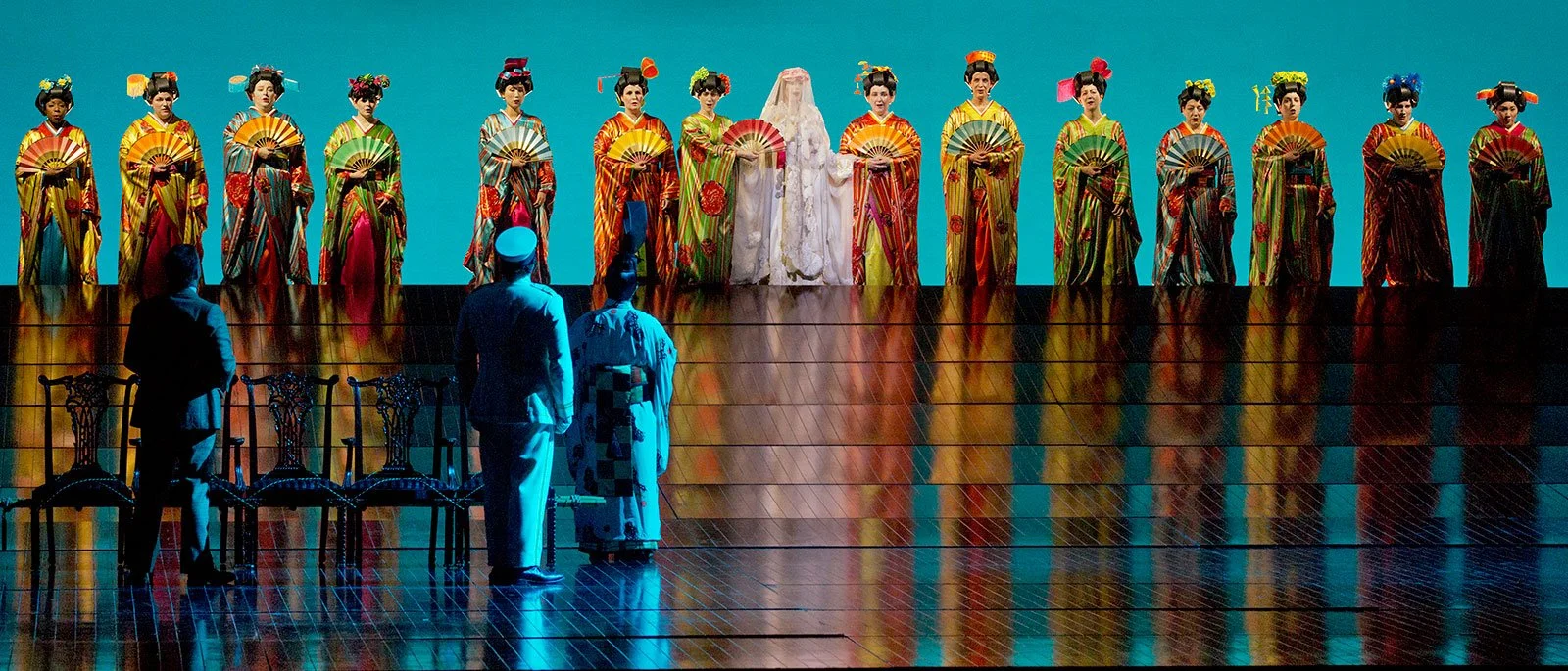 The image shows a stage with twelve women dressed in colorful traditional kimonos and umbrellas, standing in a line. In the foreground, three men in different costumes are observing the display.