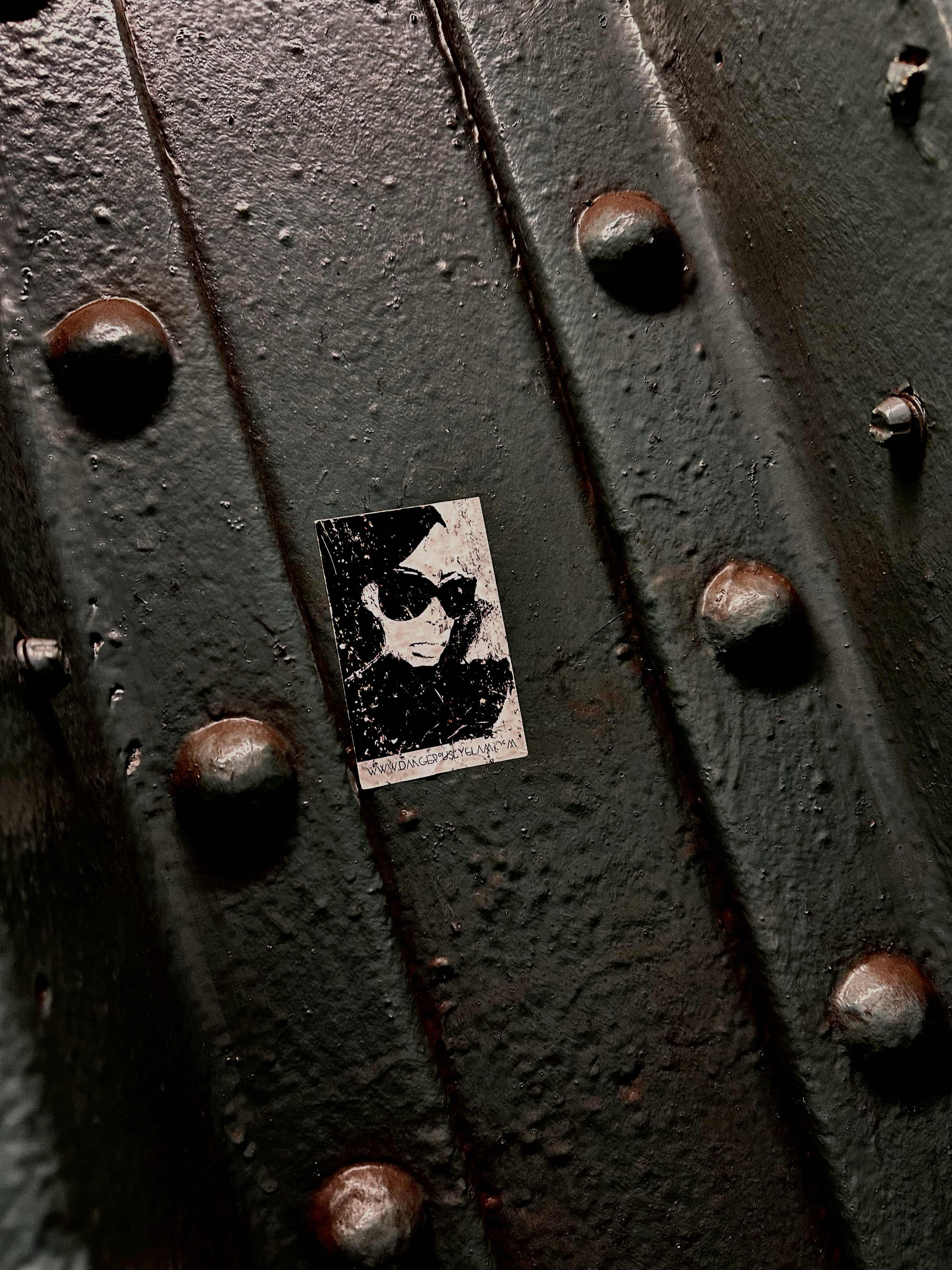 Close-up of a black, riveted metal surface with a small sticker featuring a black and white illustration of a woman wearing sunglasses and a leather jacket.