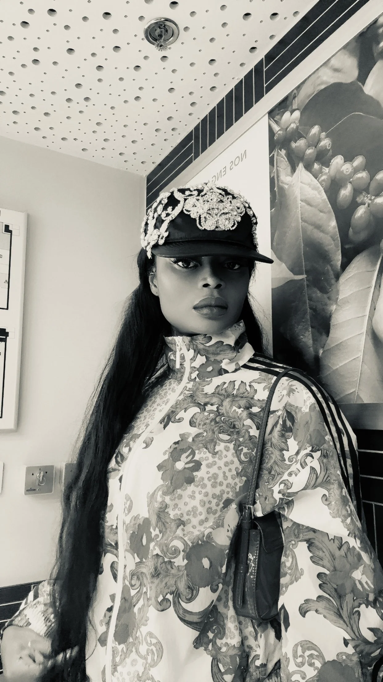 A woman wearing a patterned jacket, a cap with decorative embellishments, and carrying a small bag. She has long dark hair and is standing indoors near a wall with large leaf images.